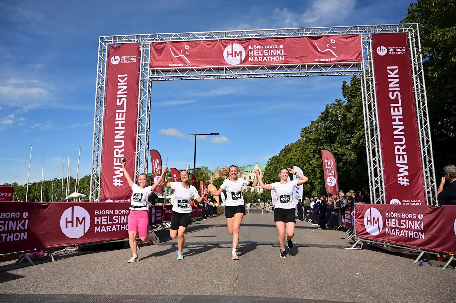 This image shows a group of people crossing the finish line of the Helsinki Marathon. They are celebrating as they complete the race. The banner and signage indicate the event's sponsor and location. The weather appears to be clear, and there are spectators around the area.