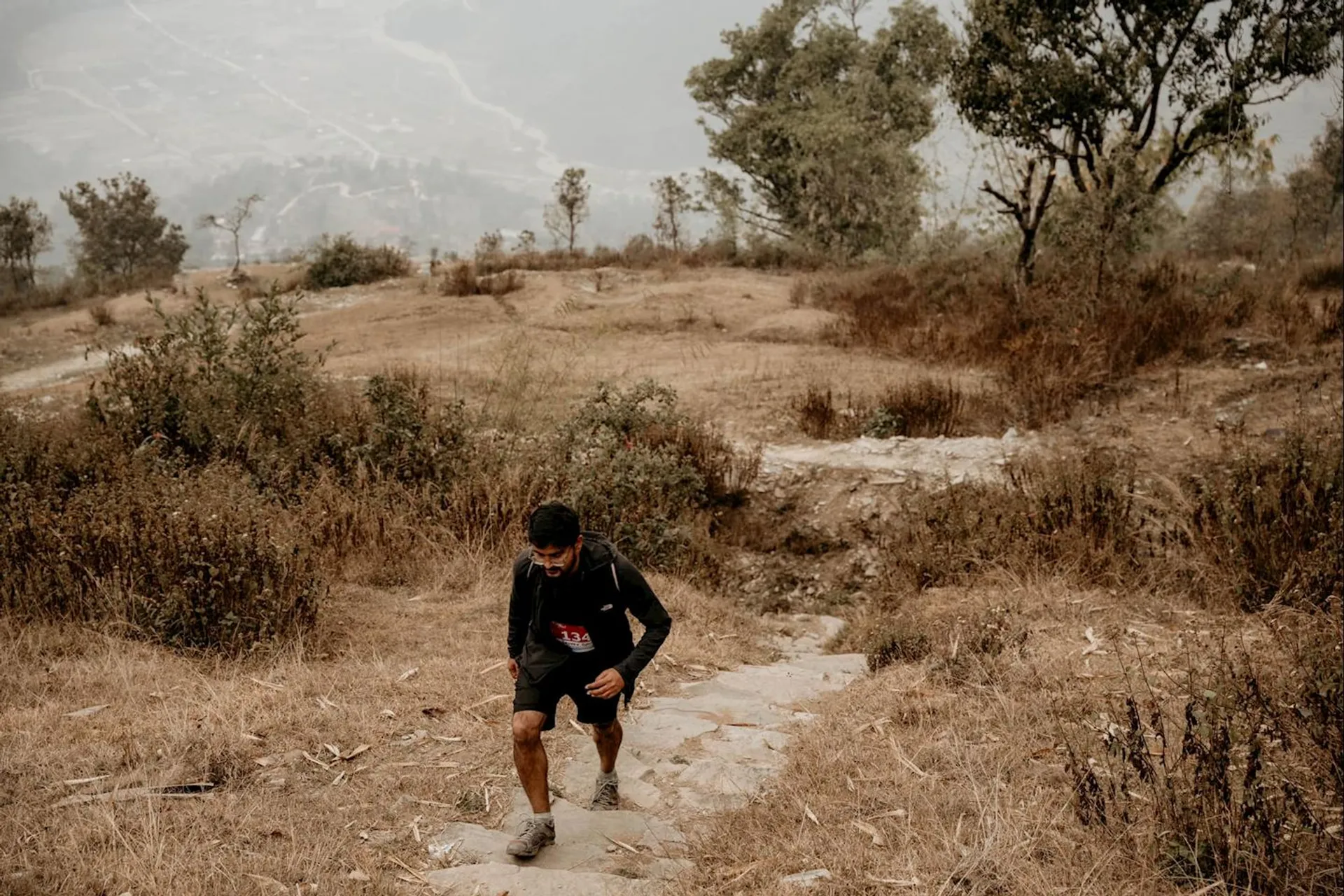 The image shows a person hiking up a rocky trail on a hillside. The surrounding area is dry with grass and some shrubs, and there are trees in the background. It appears to be an outdoor, natural setting with a distant view of a valley or landscape below.