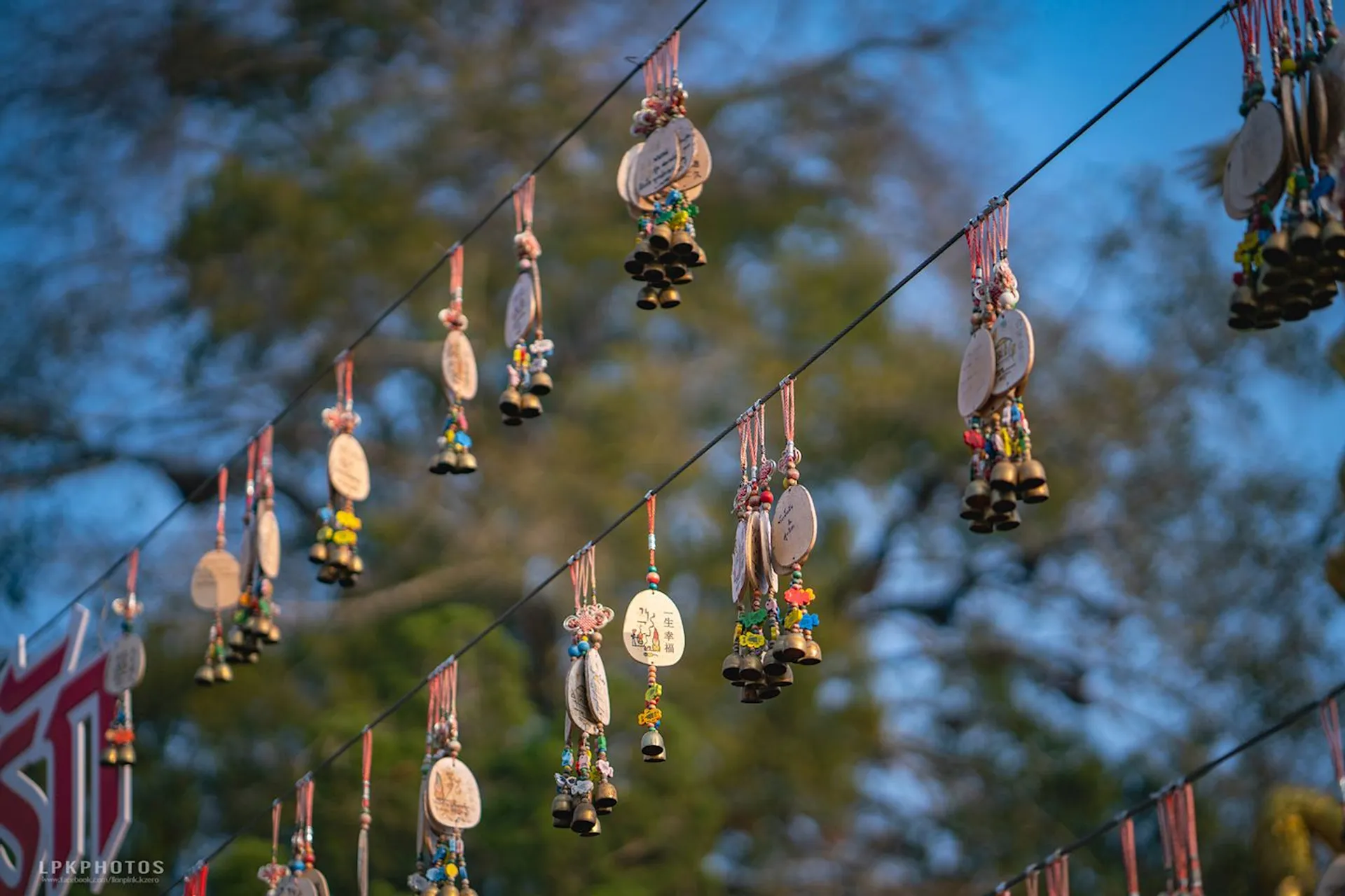 The image shows several decorative hanging objects suspended on strings. These objects appear to be small, colorful charms or ornaments, possibly with wooden or metal components. They are artistically strung along the wire, with trees and a clear sky in the background, suggesting they are displayed outdoors.