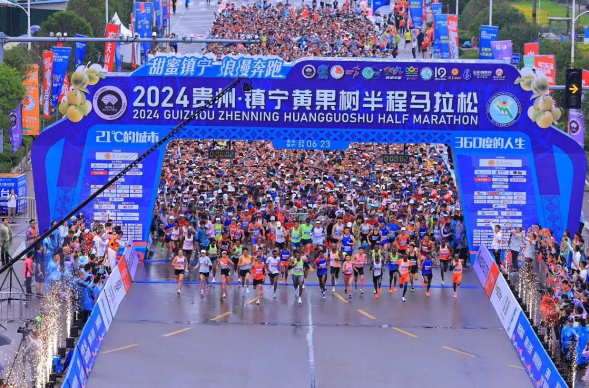This image shows participants at the start line of the 2024 Guizhou Zhenning Huangguoshu Half Marathon. The event banner is prominent at the top, and a large crowd of runners is seen preparing to begin the race. The scene is festive, with decorations and signs in the surroundings.