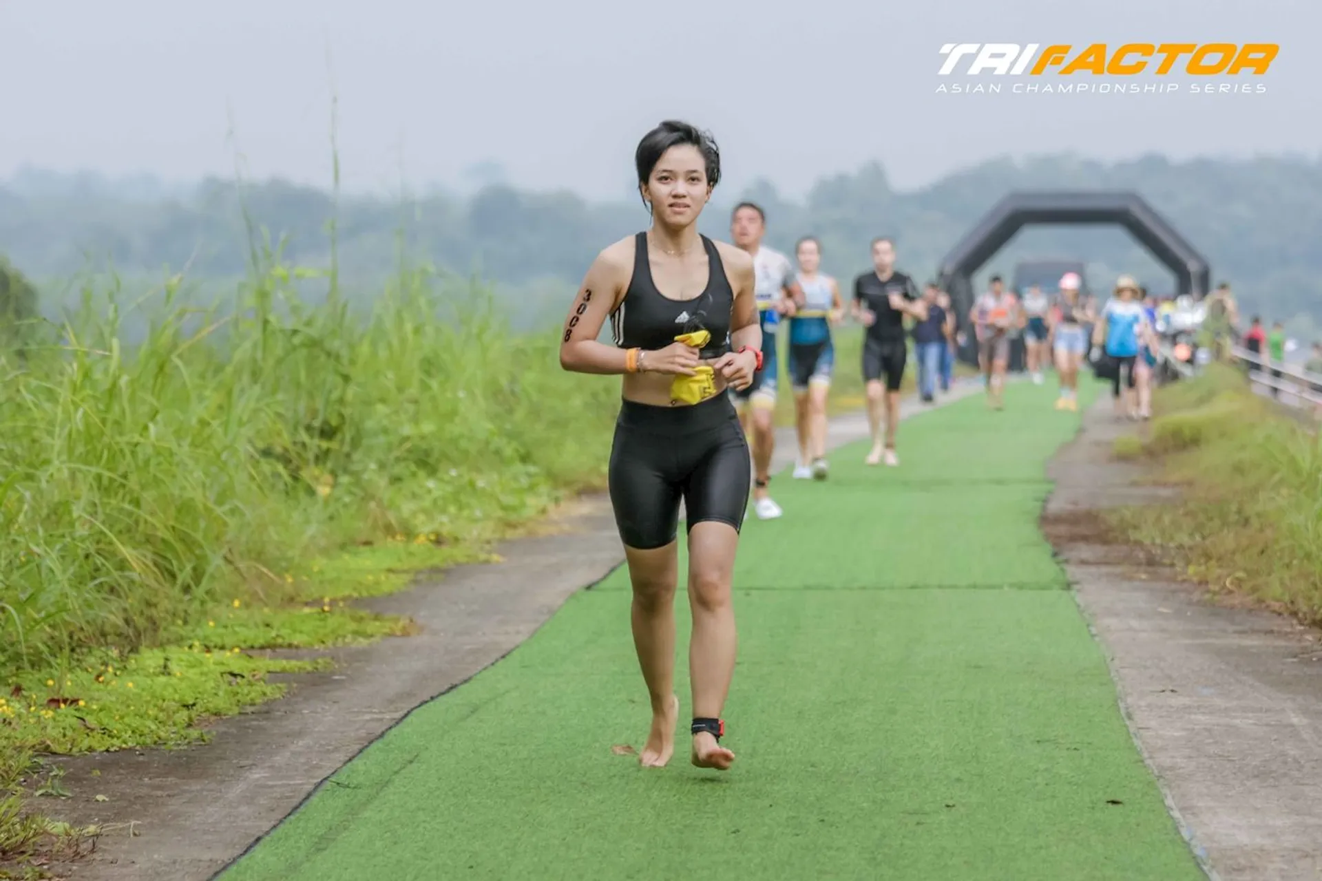 The image shows a person participating in a triathlon event, running on a green path. The event appears to be part of the TriFactor Asian Championship Series. There are other participants in the background, and the surrounding environment is natural and outdoor.