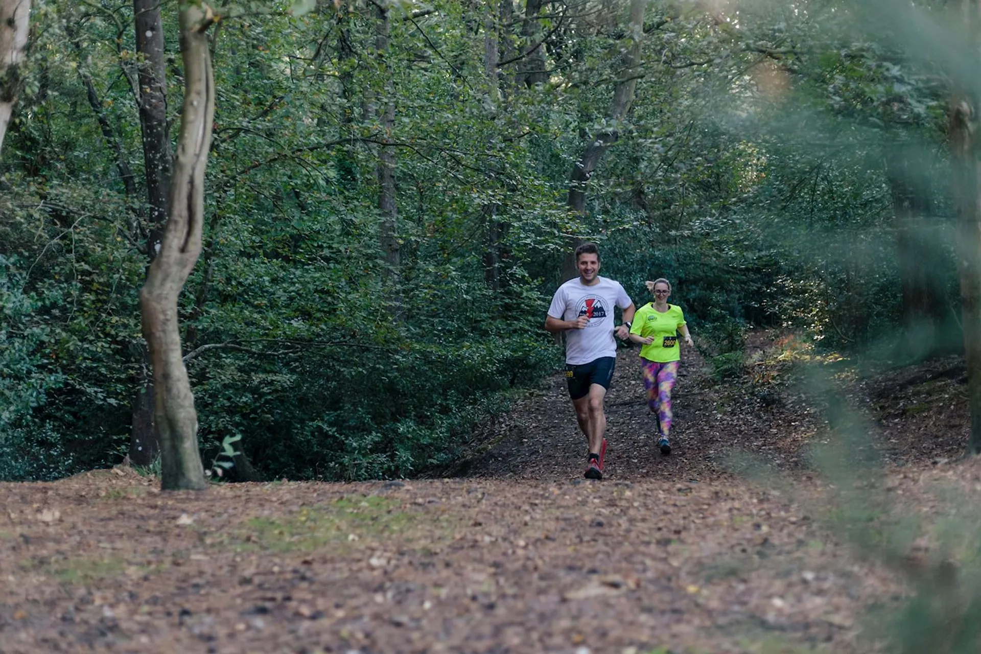 The image shows two individuals jogging or running on a dirt path through a wooded area