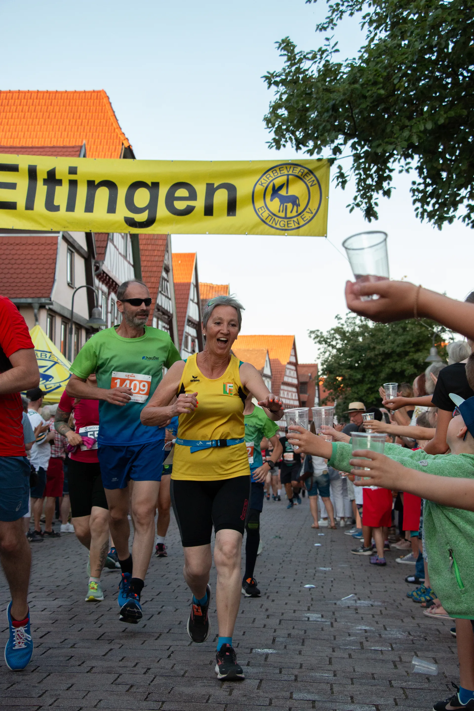 The image shows a group of runners participating in a race. They are passing by a water station where people are handing out cups of water. There is a banner with the word "Eltingen" visible above them. The scene is lively with spectators on the side and typical town buildings in the background.