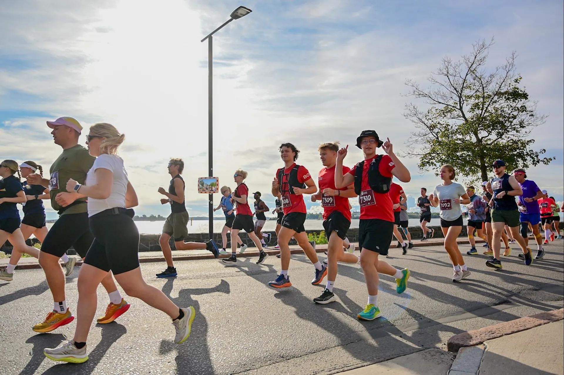 The image shows a group of people participating in a road race or marathon. The runners are on a paved path near a waterfront. They are wearing athletic clothing and have race numbers attached to their shirts. The weather appears to be clear, and a few trees and a light pole are visible in the background.