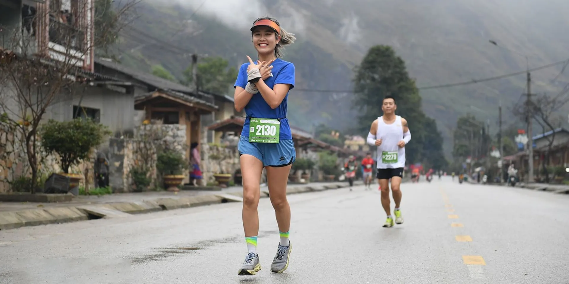 The image shows people running on a road, likely participating in a marathon or race. The person in the foreground is wearing a blue outfit, a race bib, and appears to be smiling. Other runners are visible in the background. The setting seems to be a rural area with houses along the road and hills or mountains in the background.