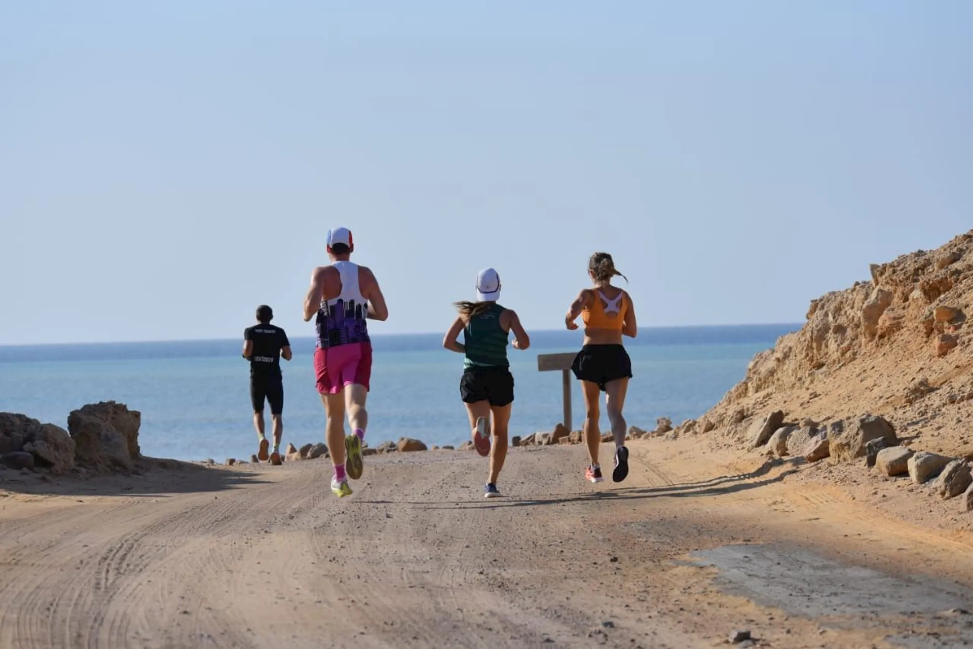 The image shows four people running on a dirt path along a coastal area. They're heading towards the sea, and the path is flanked by rocky terrain. The sky is clear, and the overall scenery appears to be sunny and warm.