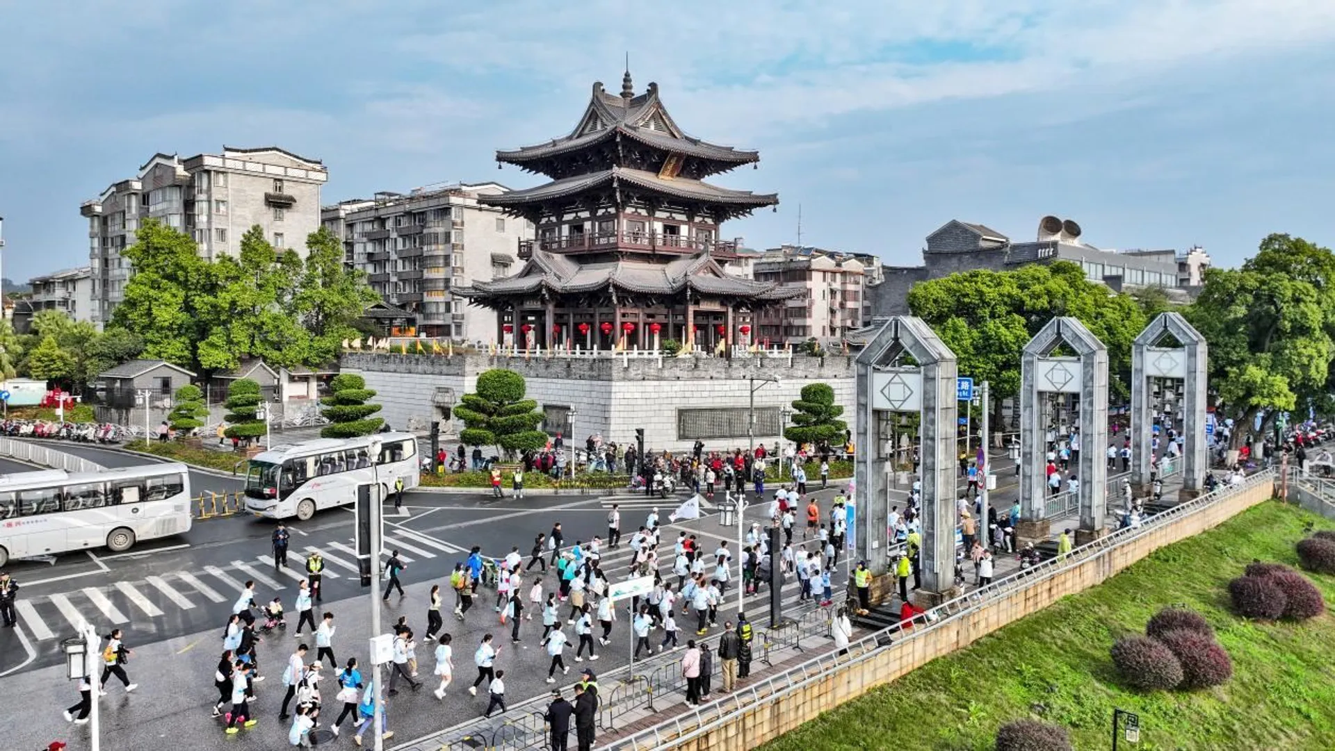 The image depicts a bustling street scene with a marathon or large group event in progress. Participants, wearing similar shirts, are running along a road. On the left, there are buses and vehicles on a crossroad. In the background, there's a traditional multi-tiered pagoda-style building surrounded by other modern structures. The area is lively and seems to be part of an urban setting with greenery around.