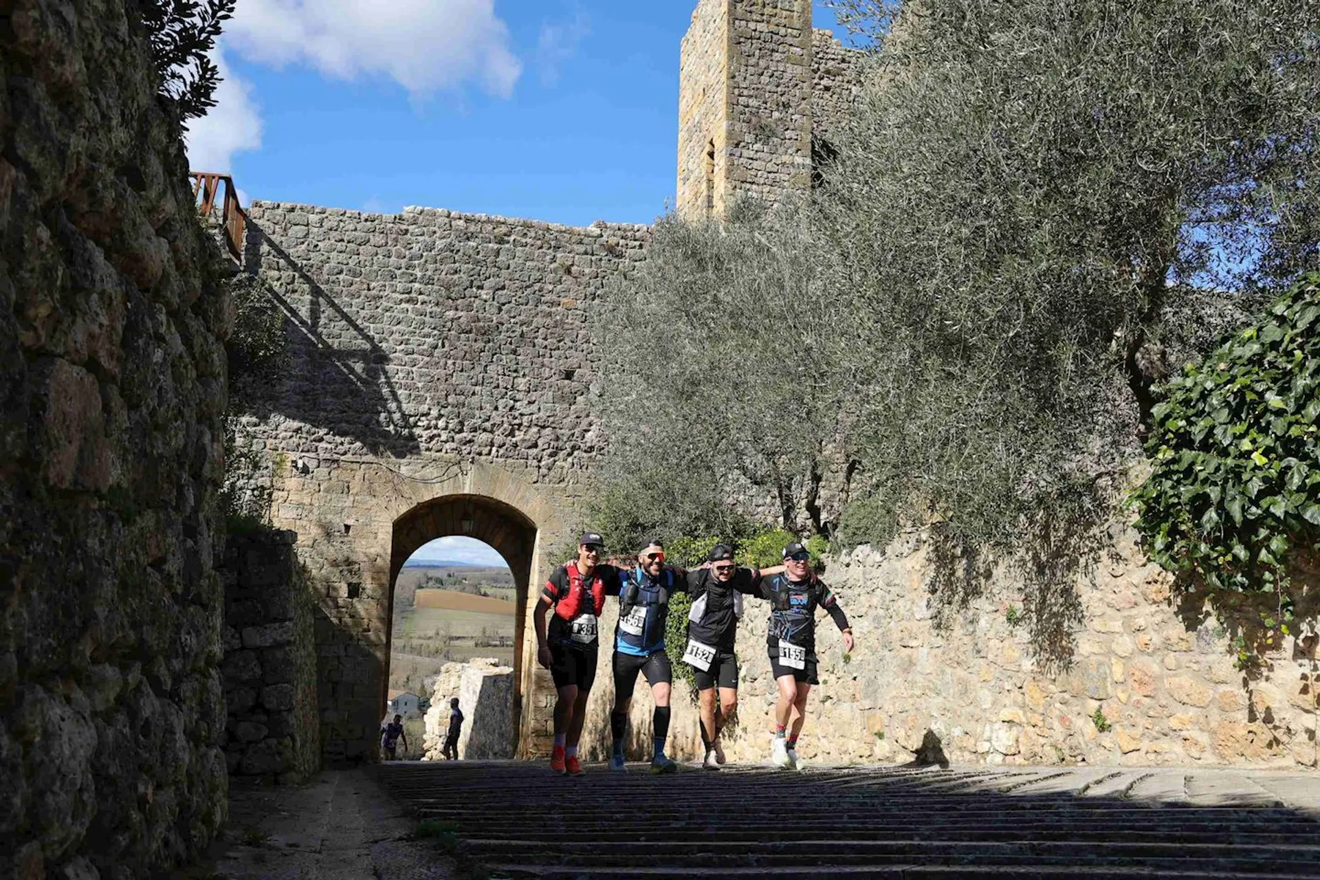 The image shows a group of four individuals, likely participating in some sort of running or trekking event, as they are dressed in athletic gear, including shorts, running shoes, and hydration packs. They appear to be running together through a historic location, indicated by the old stone architecture around them. There is a large stone wall, a tower-like structure, and an archway that they are just about to pass under. The setting is quite picturesque with trees, clear skies, and old stone pavements adding to the charm of the scene.