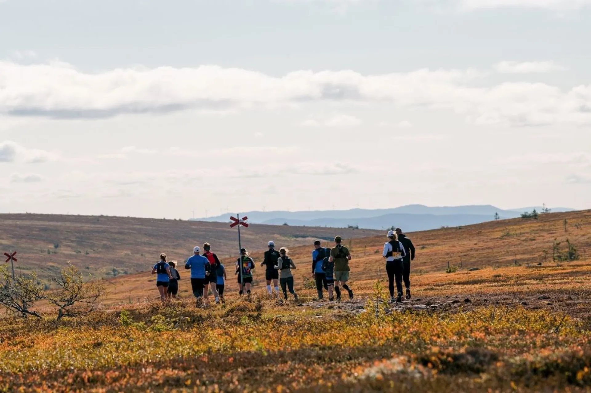 The image shows a group of people running or hiking along a trail in an open, expansive landscape. The terrain appears to be a mix of grass and low vegetation, with distant hills or mountains in the background. The sky is partly cloudy, and the setting looks like it could be a remote or wilderness area. There's a signpost marker visible on the trail.