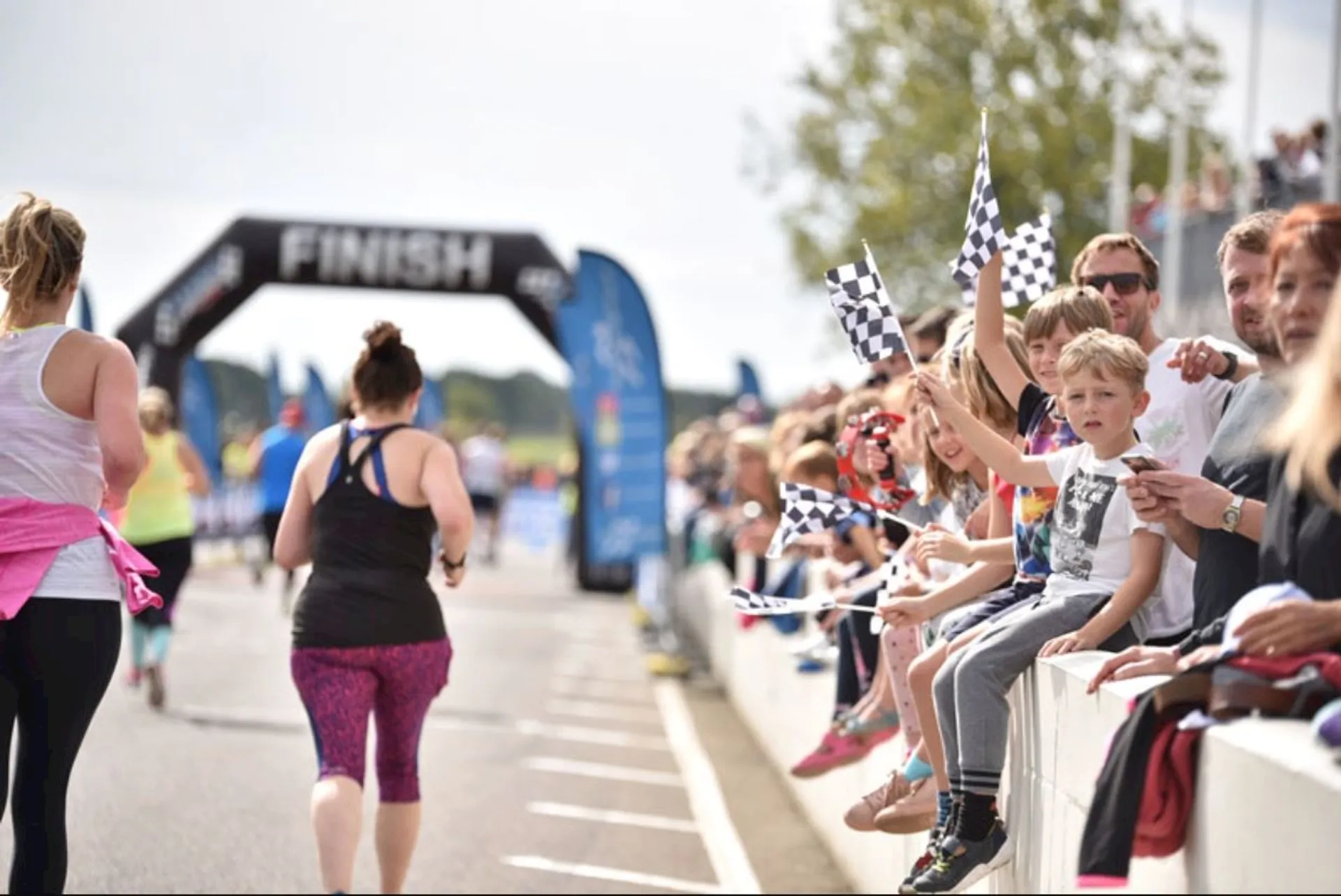 The image shows a running event with participants heading toward a "FINISH" line. On both sides of the path, there are spectators, some of whom are sitting on barriers and waving checkered flags. The scene is lively and appears to be taking place outdoors on a sunny day.