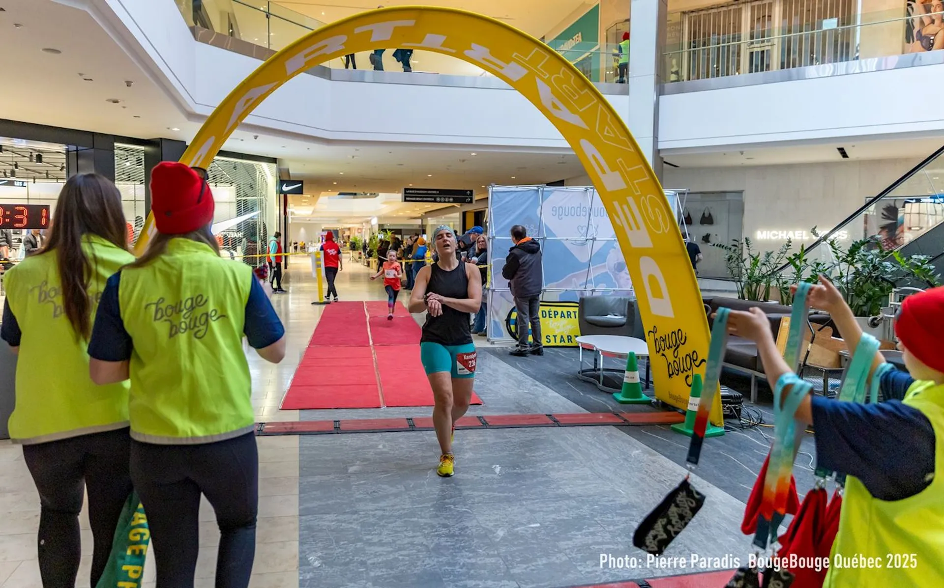 In the image, a person is running indoors through a finish line marked by an archway. They appear to be participating in an event, possibly a race or marathon. There are other people in bright yellow vests watching and guiding the runner. The setting looks like a shopping mall, with multiple floors visible.