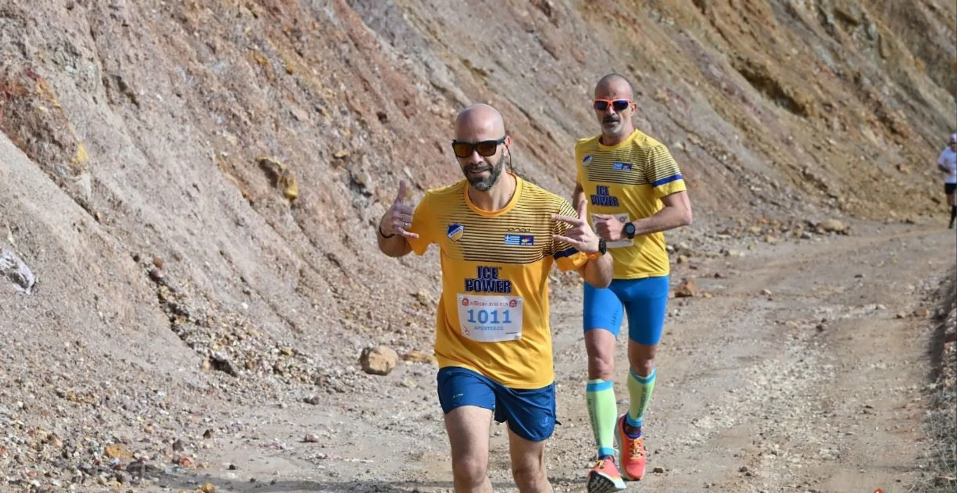 The image shows two people running on a rough trail, likely in a race or marathon. They are wearing yellow shirts, and one runner has a visible race bib with the number 1011, as well as sunglasses. The background features a rocky, uneven terrain.