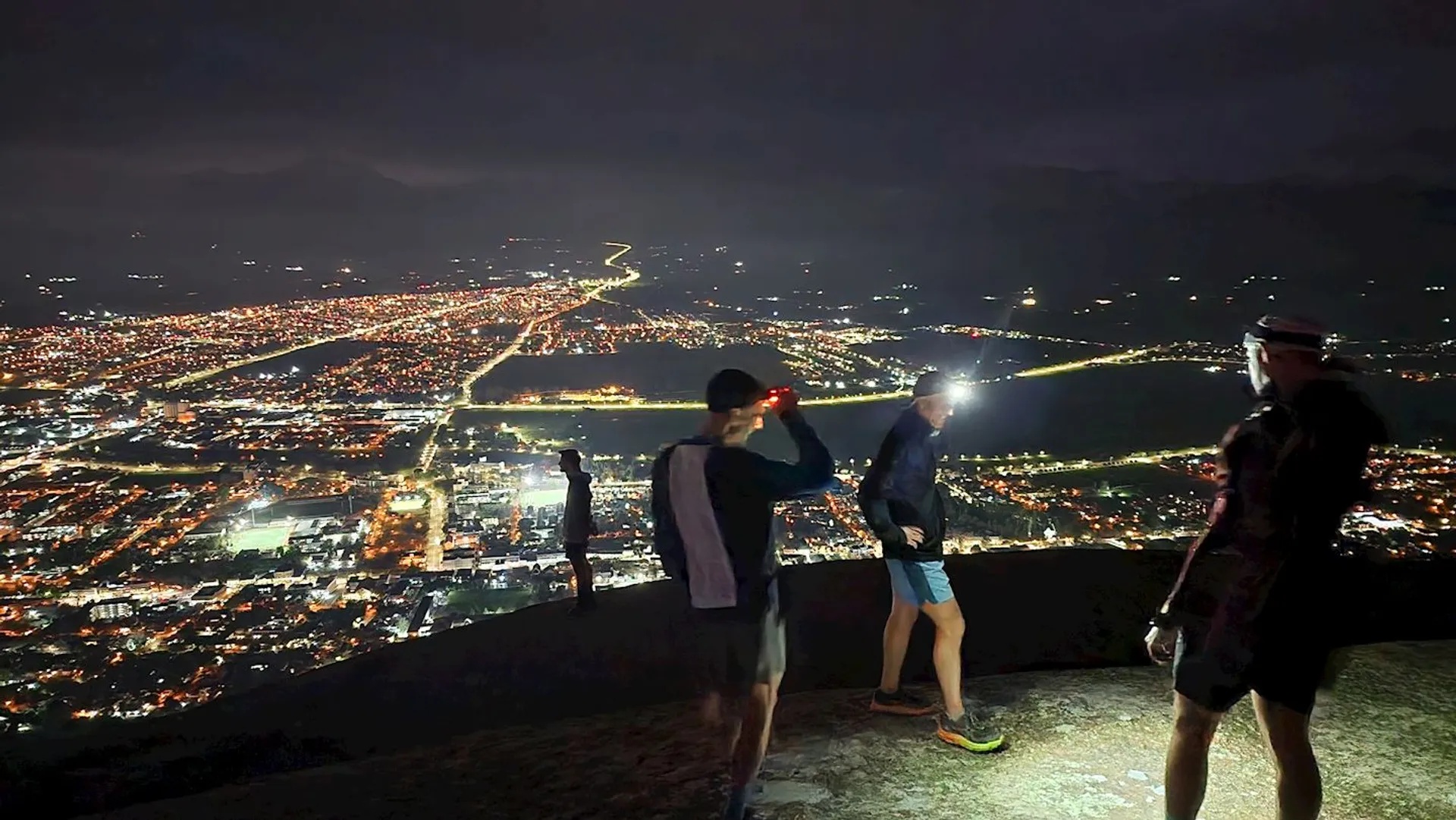 This is an image of several individuals standing on an elevated viewpoint at night, overlooking a cityscape below. The city is illuminated with numerous lights, outlining streets and defining various areas of urban development. The people are seemingly enjoying the view, with one person gesturing towards the city. It appears to be an outing or a hike that they've taken to reach this vantage point. One of them is using a flashlight to either light the path or point towards something specific in the distance. The atmosphere suggests a sense of adventure and appreciation for the nighttime city view.