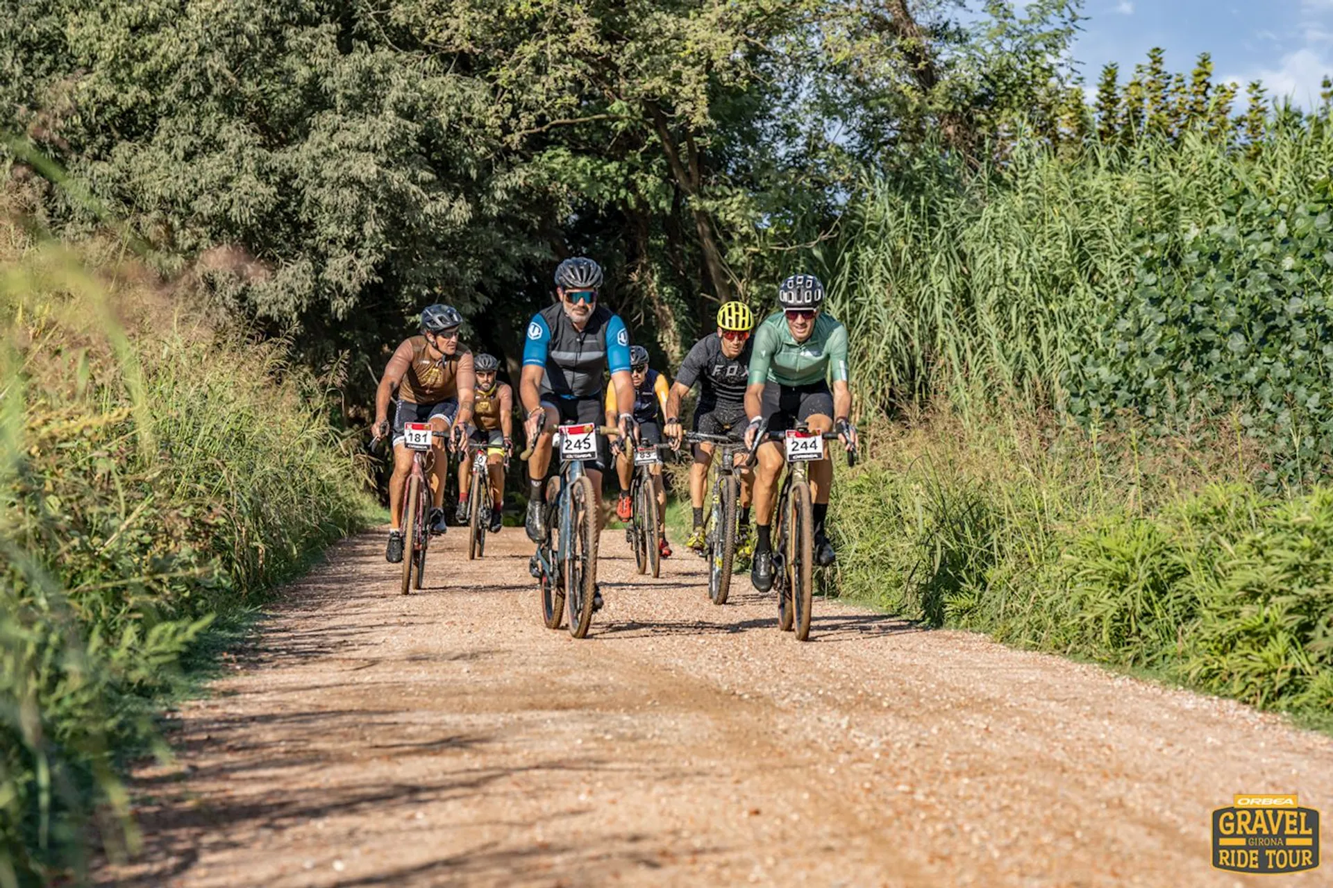 The image shows a group of cyclists riding on a gravel trail outdoors. They appear