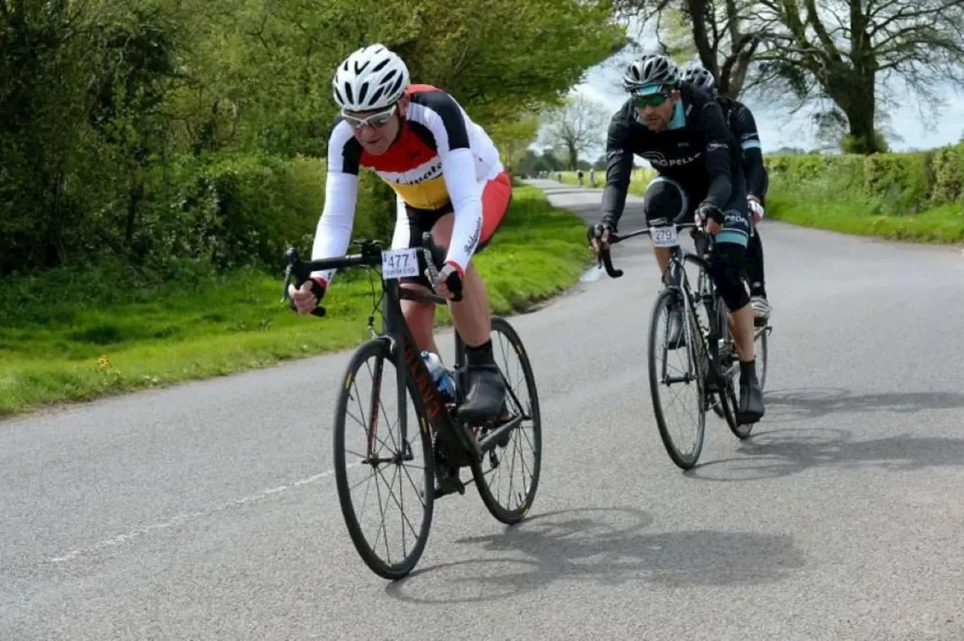 The image shows two cyclists in a road cycling race or event. They are dressed in cycling gear, with the foremost cyclist wearing a white helmet, a white jersey with red and yellow accents, and black shorts, while the following cyclist is dressed in black with a black helmet. Both are riding road bikes, which are characterized by their slender tires, lightweight frames, and dropped handlebars for a more aerodynamic position. The scene takes place on a road lined with green grass and trees, suggesting a rural or semi-rural environment. The cyclists appear to be exerting effort, indicating they might be in a competitive or challenging part of the race.