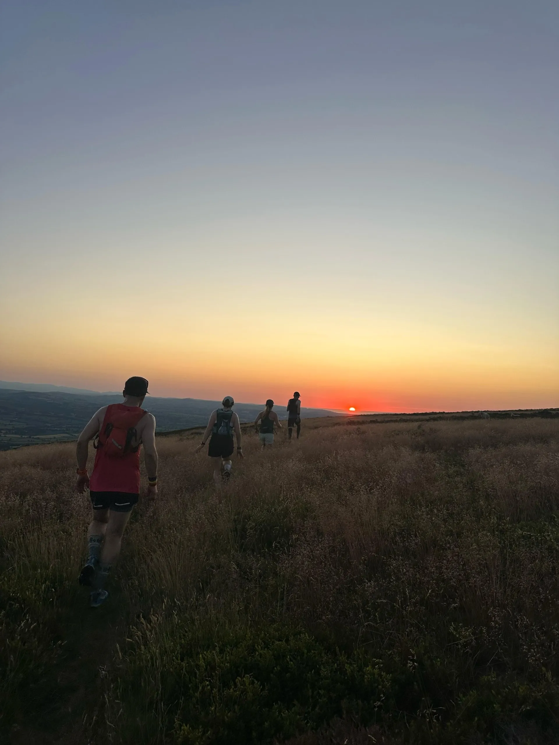 The image shows a group of people walking on a grassy path towards a sunset. The scene is outdoors, with an expansive landscape and a clear sky. The sun is near the horizon, creating a warm and serene ambiance.