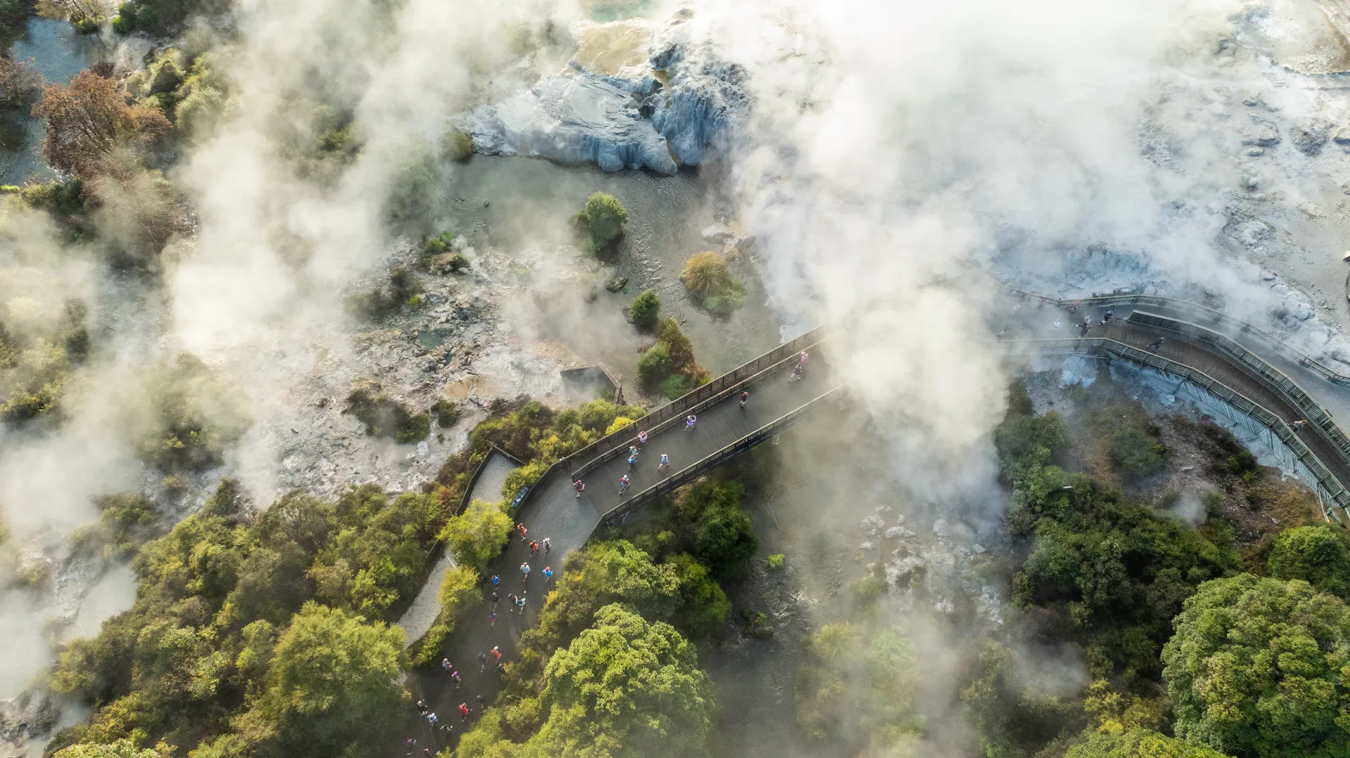 This image shows an aerial view of a winding path or boardwalk surrounded by trees and vegetation, with a misty or foggy atmosphere. There are people walking along the path, and the setting appears to be a natural area, possibly near geothermal features or hot springs, given the presence of steam and rocky terrain.