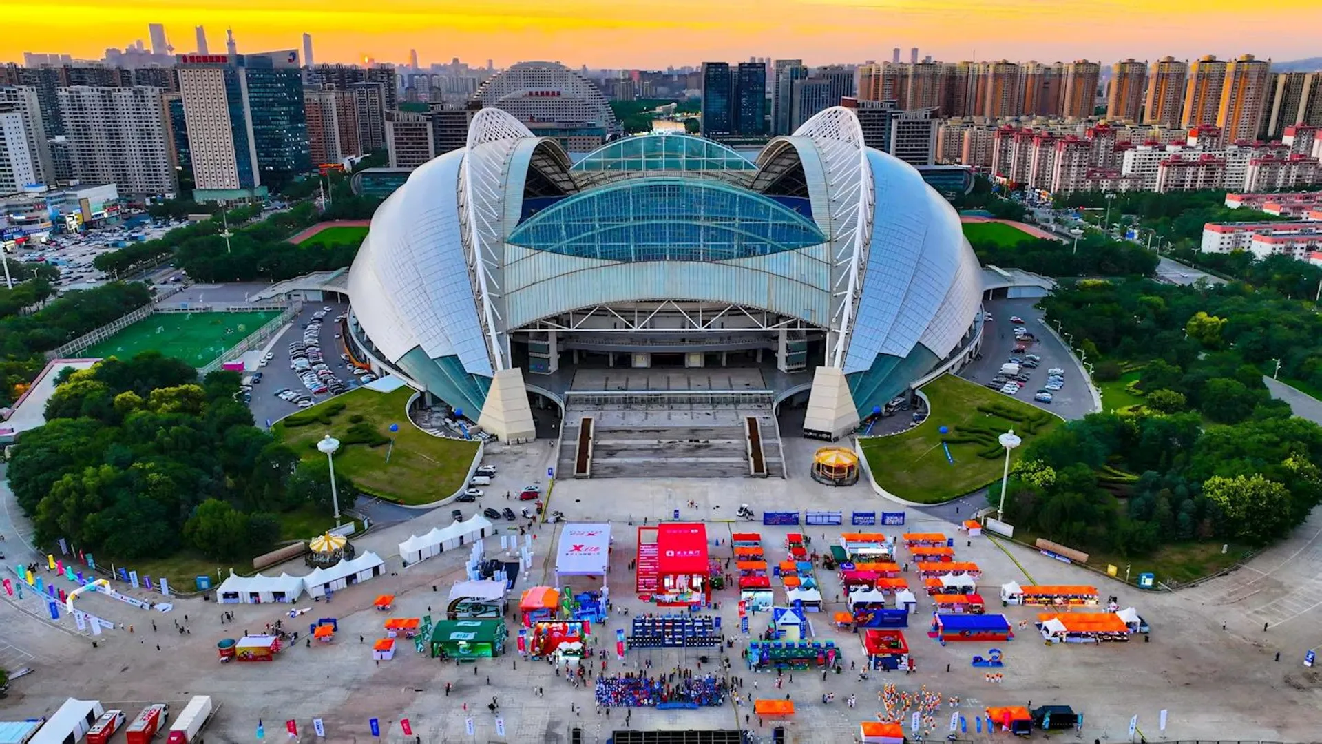This image shows a large, modern stadium with a distinctive, curved architectural design. In front of the stadium, there's an open area with numerous colorful tents and booths set up, possibly for an event or festival. Surrounding the stadium are urban buildings and greenery, and the scene is set at sunset or sunrise, giving the sky a vibrant orange hue.