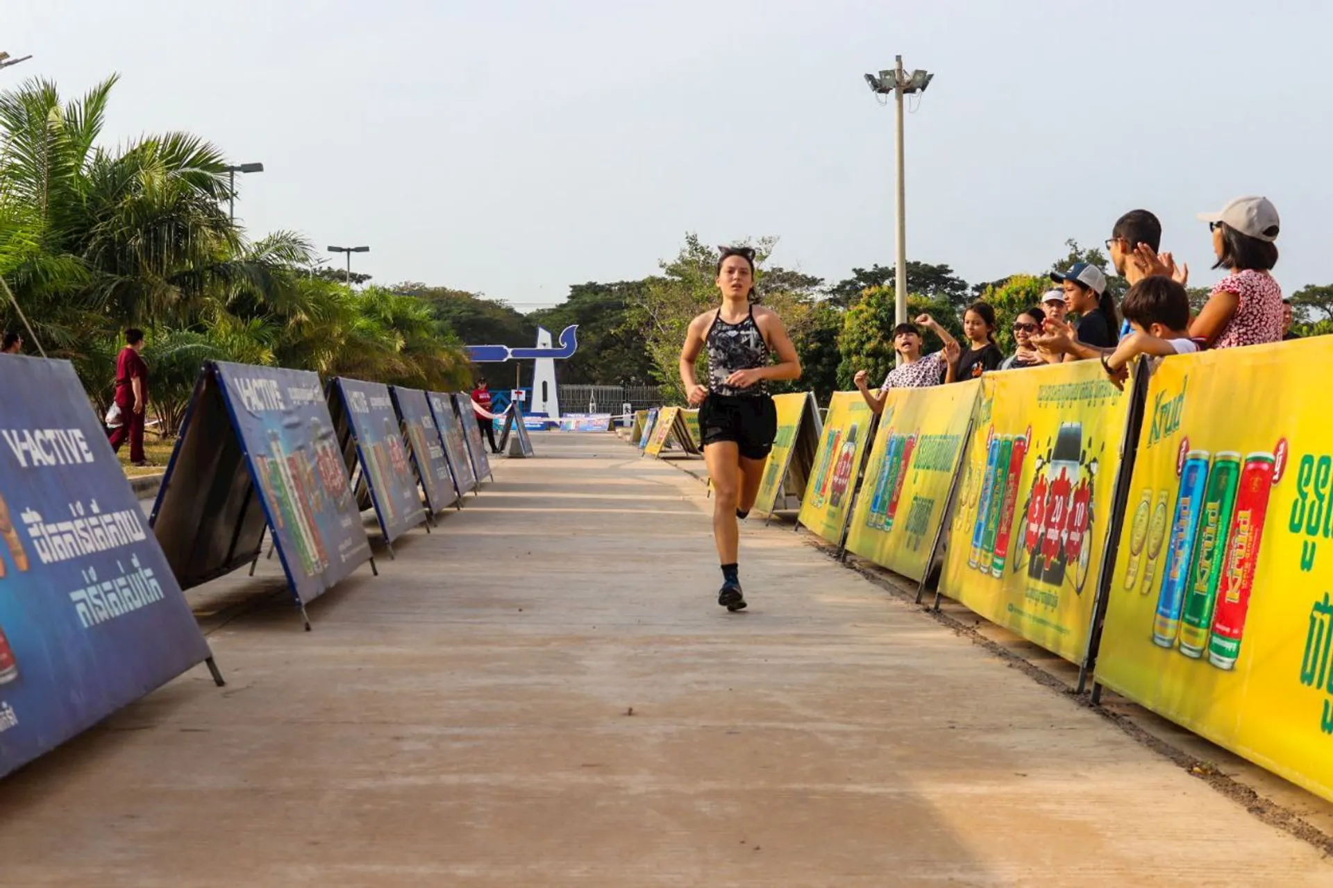 The image shows a person running on a concrete pathway, surrounded by colorful advertising banners on both sides. There are spectators on the right side of the path, watching and cheering. The setting seems to be an outdoor event, possibly a race or marathon, with trees and a clear sky in the background.