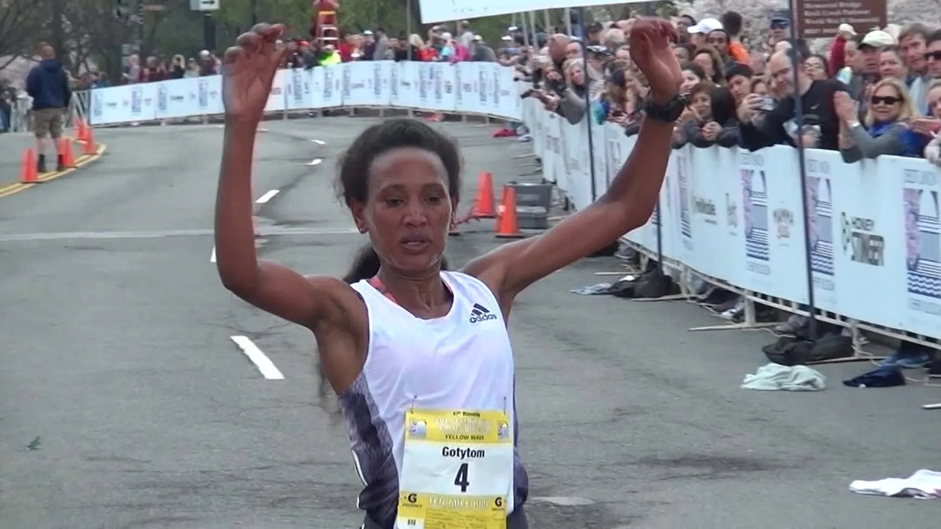 The image shows a female runner crossing the finish line at a road race. She