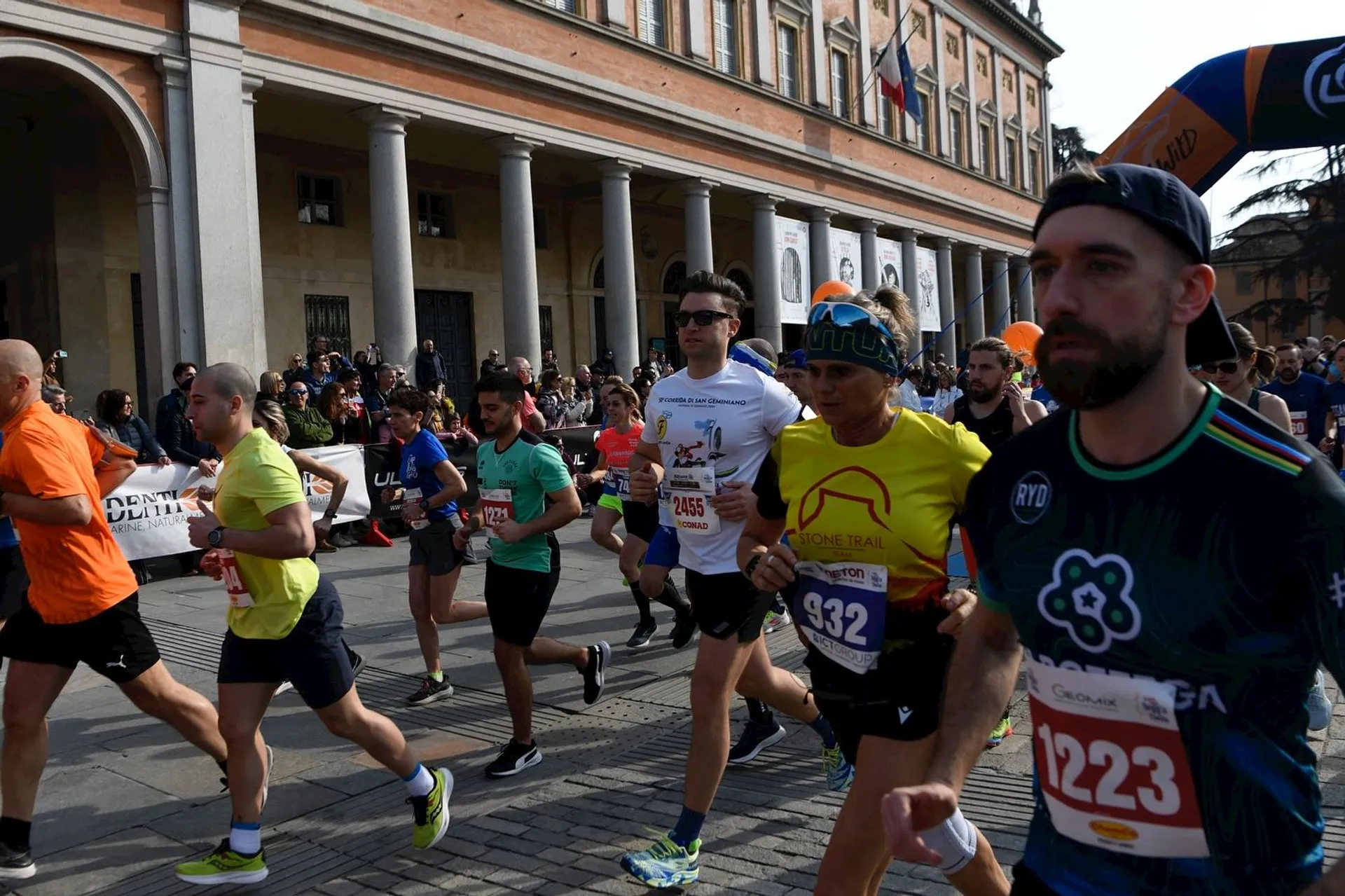 The image shows a group of people participating in a running event. They are running on a street beside a large building with columns. There are spectators and banners in the background along the sides of the street. The participants are wearing numbered bibs and athletic clothing.