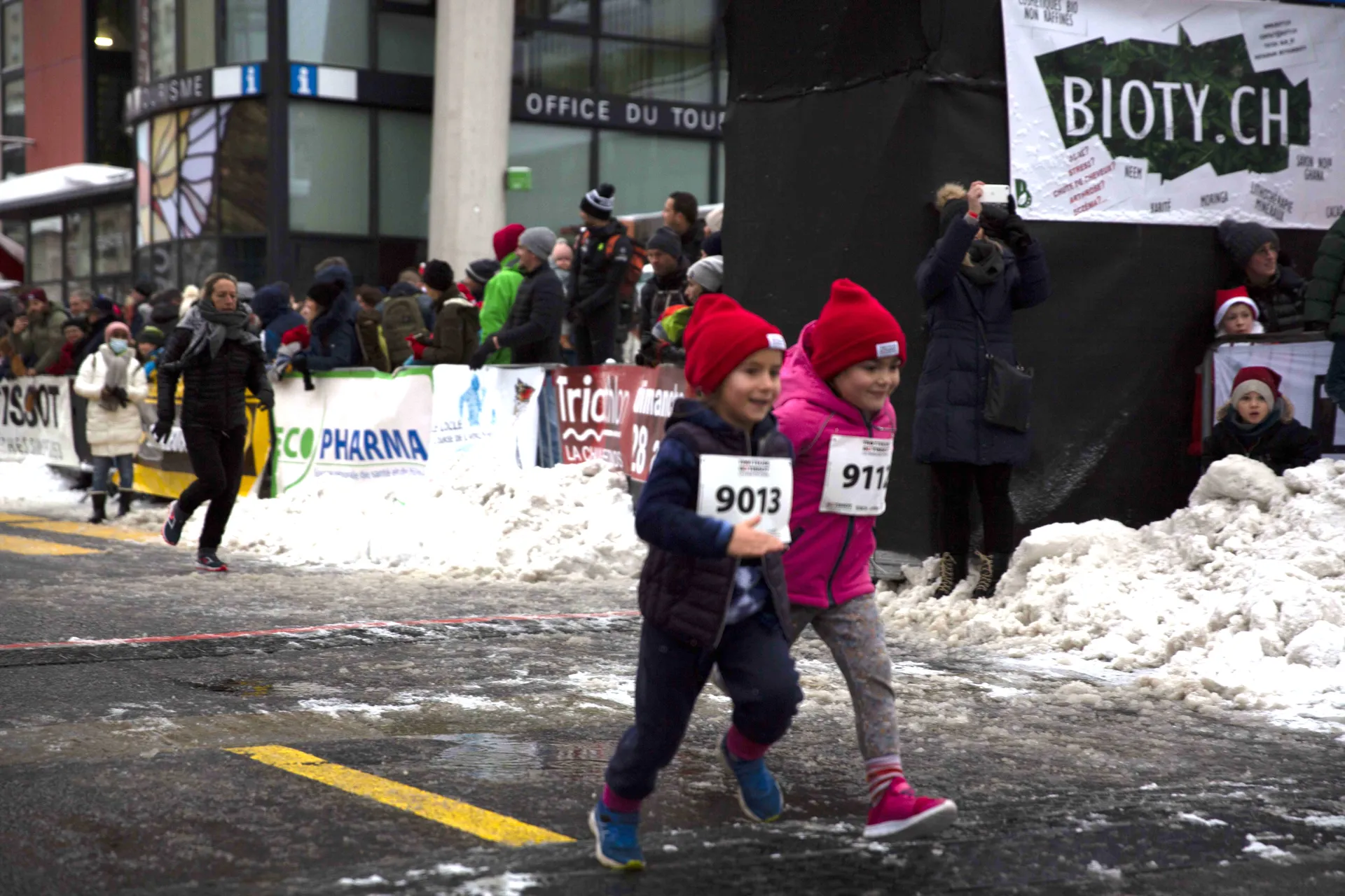 The image shows two children wearing race bibs, running on a snowy street. They appear to be participating in a running event, as indicated by the bib numbers (9013 and 911). Spectators are gathered along the sides, watching the event. The setting is wintry, with snow on the ground.
