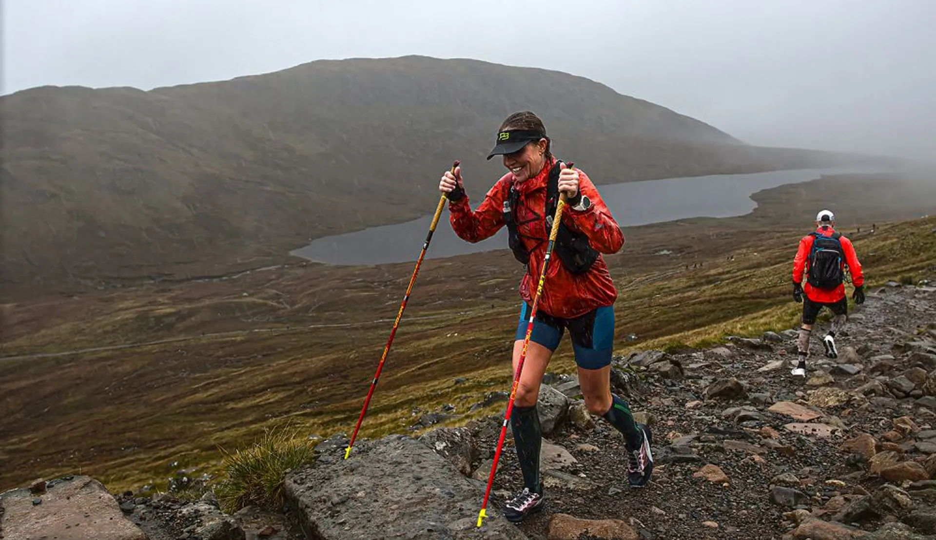 In the image, there are two individuals engaged in an outdoor activity, likely hiking or trail running. The person in the foreground is using trekking poles to aid their movement and is wearing a short-sleeve top, shorts, and a cap, suggesting they are exerting themselves physically and dressed for active wear despite the overcast weather. They seem to be focused and pushing forward at a good pace. The second person is in the background, also equipped with a backpack like the first individual, following along the same path. The terrain appears to be a mountainous landscape with rocky ground and a calm lake or loch visible in the valley below, partly shrouded by mist or low clouds, indicating that the location might be subject to changeable