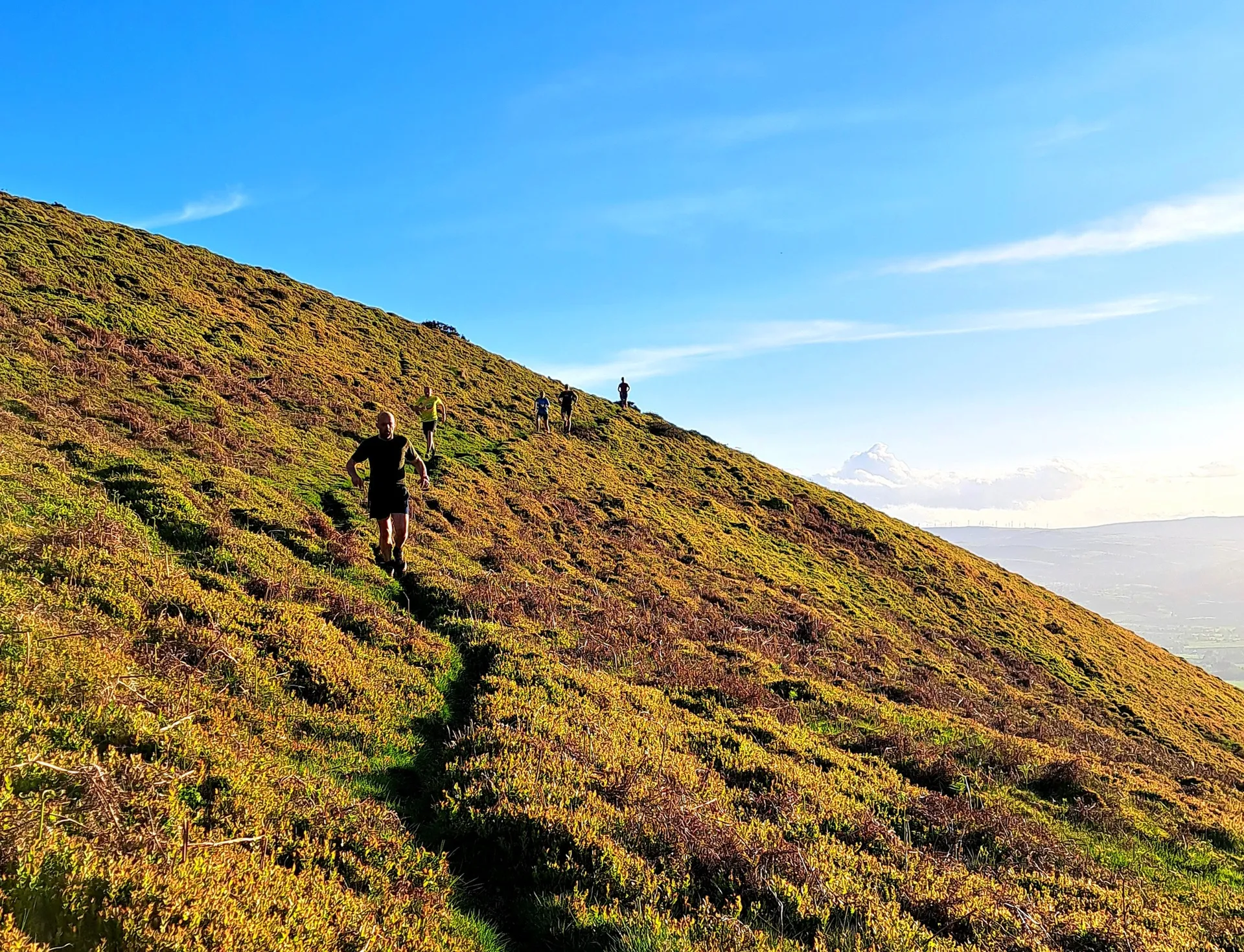 The image shows a scenic landscape with several people walking or jogging up a grassy hillside. The sky is clear and blue, and there are distant mountains visible under the clouds, creating a picturesque view. The hill is covered with vegetation, and the terrain appears to be a natural trail.