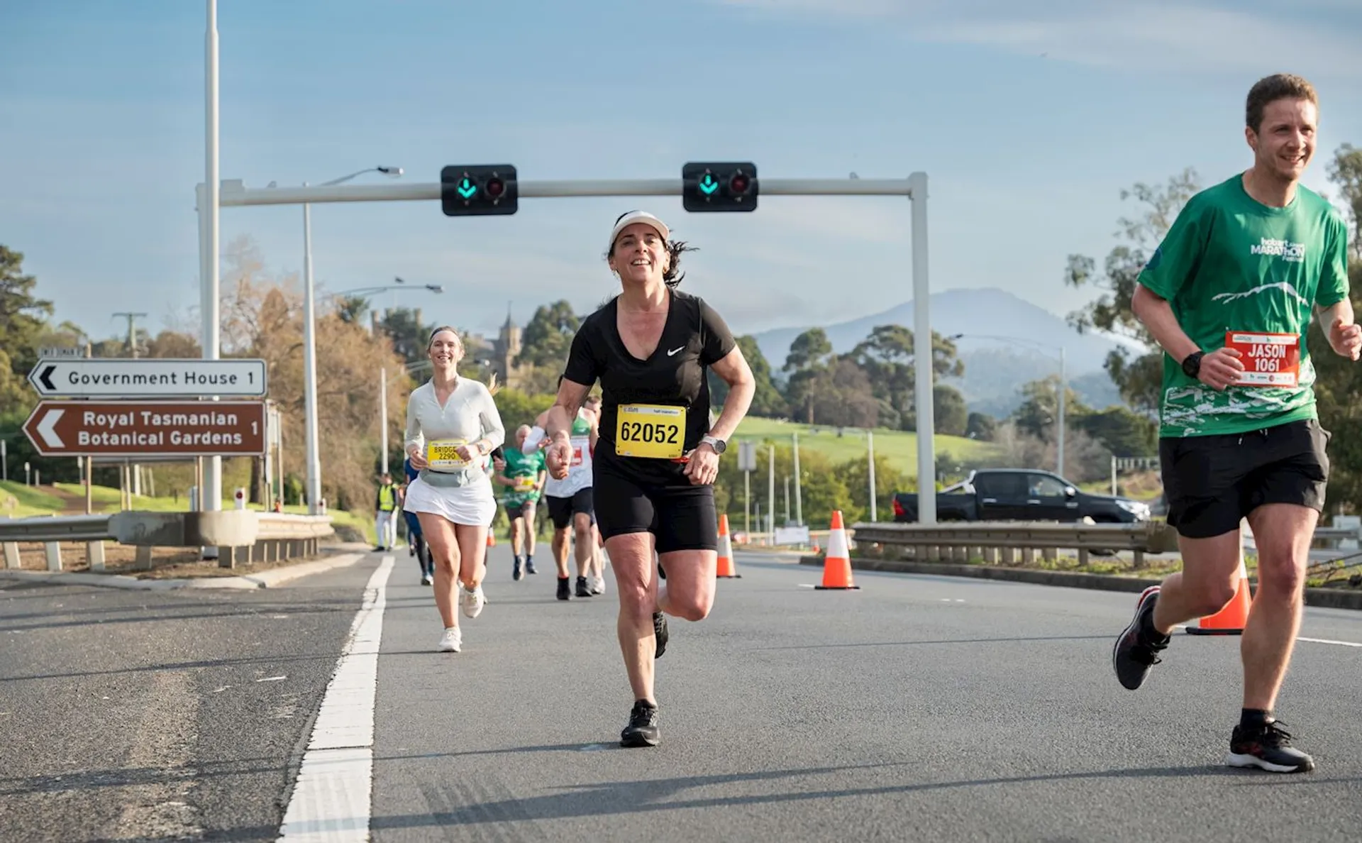 The image shows a group of people running on a street, possibly participating in a race or marathon. They are wearing athletic clothing and bibs with numbers. There are traffic cones and a sign indicating directions, with a scenic background of greenery and hills.