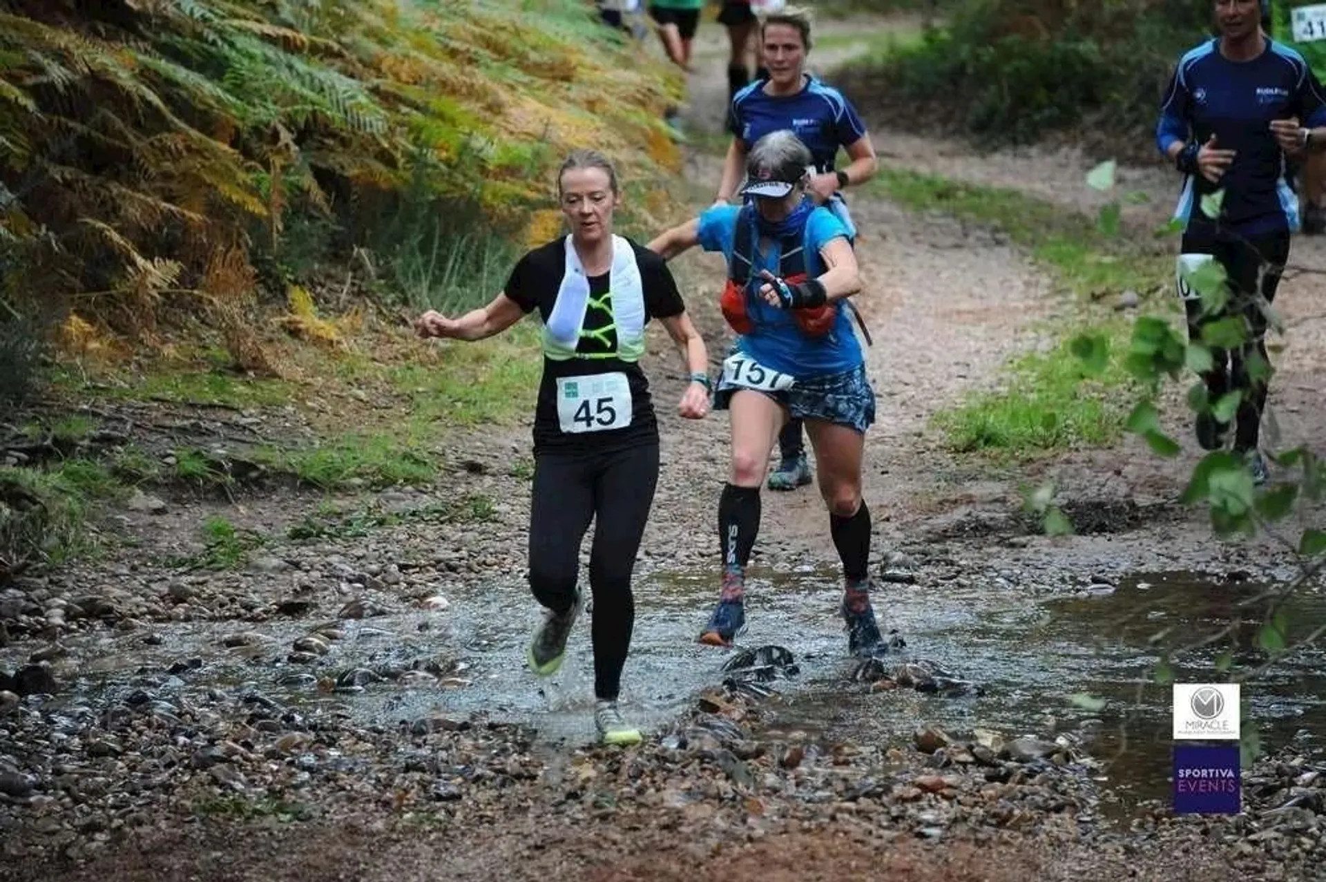 The image shows two people participating in a trail run, splashing through a shallow