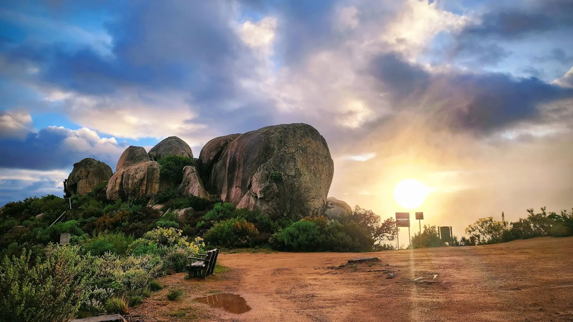 The image shows a stunning landscape scene featuring large rock formations under a sky with dramatic clouds. The sun is low in the sky, indicating it could be either sunrise or sunset, and it casts a warm glow across the scene. Vegetation, which appears to be a mix of shrubs and grasses, grows around the rocks, suggesting a natural and possibly protected area. A bench is positioned near the left side of the frame, inviting viewers to sit and enjoy the view. There is also a signpost visible on the right, but the details on the sign are not legible. The ground in the foreground looks like a dirt path or clearing. The scene is serene and picturesque, typical of a scenic viewpoint or nature reserve.