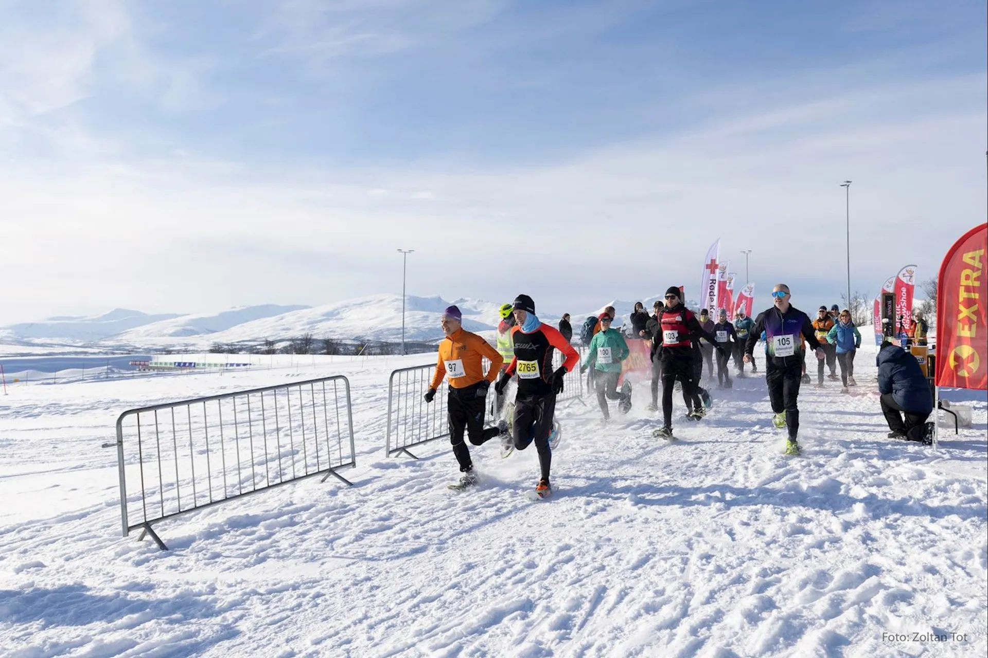 The image shows a group of runners participating in a race on a snow-covered landscape. They are dressed in athletic gear suitable for cold weather, including long-sleeve tops and running tights, and appear to be just at the starting line or early in the race, as indicated by the presence of the race barriers and the starting arch in the background. The setting includes a wide-open space with snow on the ground and distant mountains, creating a scenic but likely challenging environment for a running event. The sky is clear, suggesting good weather conditions despite the snow. There are also various race banners and flags adding a touch of color to the predominantly white landscape.