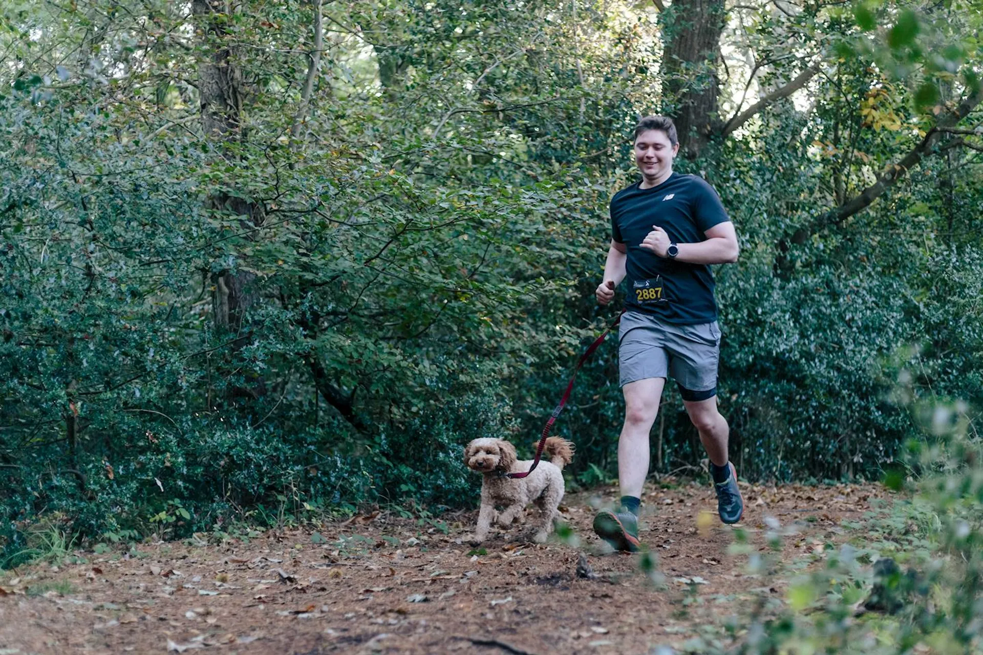The image shows a young man running through a wooded area, accompanied by a dog