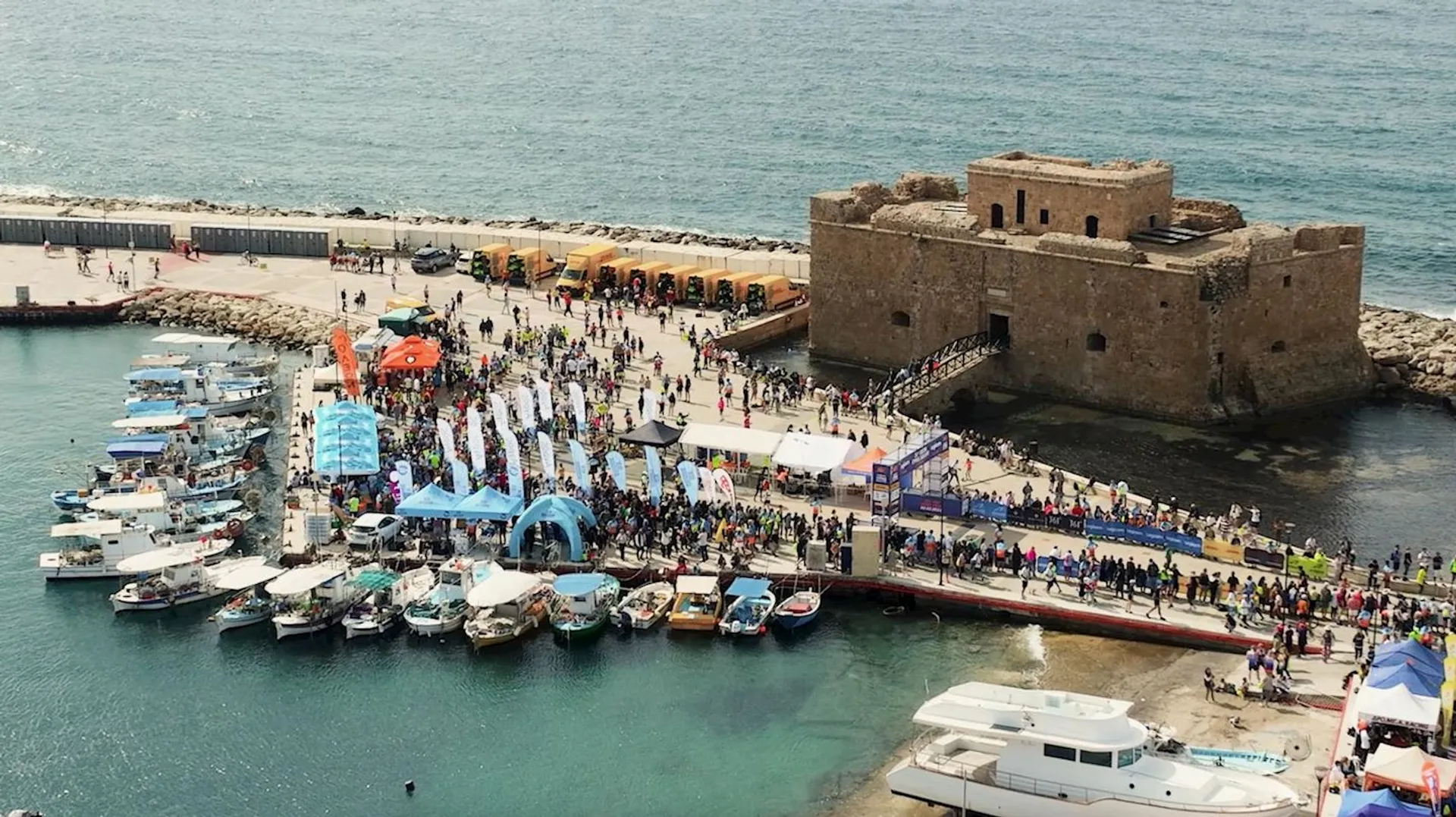 The image features a coastal scene with a large gathering of people, boats, and a prominent stone fortress on a pier extending into the sea. There are multiple tents and flags indicating an event or festival, and a variety of boats ranging from small personal watercraft to larger yachts are moored along the docks. The atmosphere seems lively, with the presence of visitors and possibly participants of a boating or maritime event.