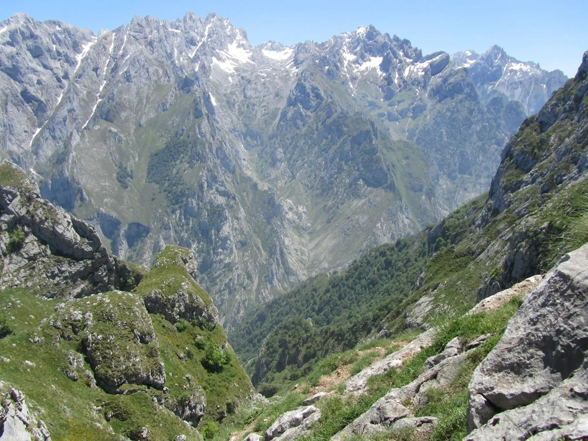 This image shows a mountainous landscape with rugged peaks, rocky terrain, and patches of greenery. There are areas of snow on the higher peaks, suggesting a high-altitude environment. The scene is likely part of a natural mountainous region, showcasing dramatic elevation changes and varied textures.