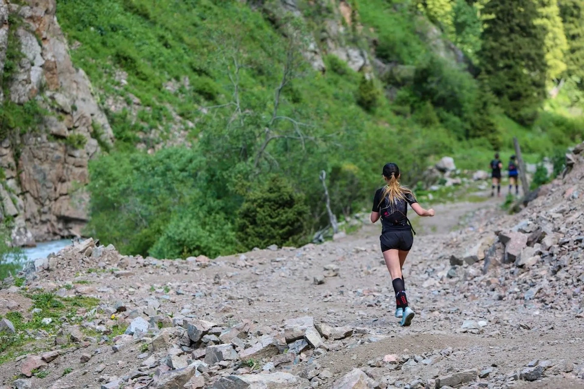 This image shows a woman running on a rocky trail in a mountainous environment.