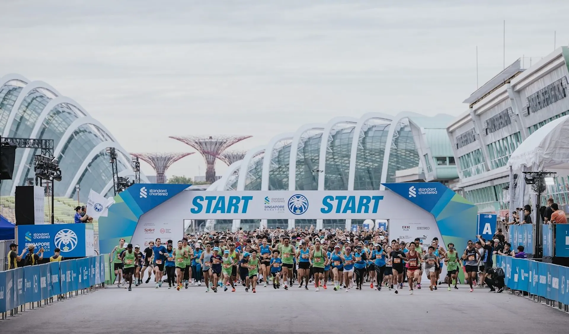 This image shows a large group of people participating in a running event. They are starting a race, indicated by the "START" banners above them. The setting includes modern architecture and structures with a crowd of runners wearing athletic attire.