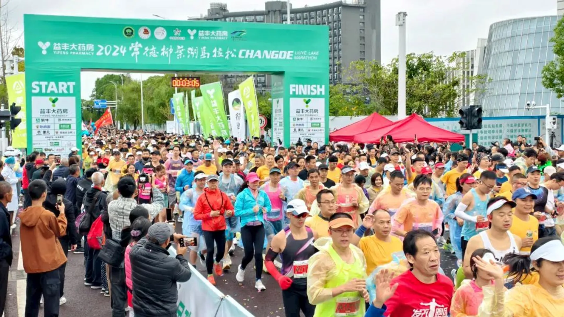 The image shows a large group of people participating in a marathon or running event. There is a starting line with banners and a clock displaying a time. Participants are wearing varied running gear, some with rain ponchos, indicating it might be rainy weather. The event is likely sponsored or themed, given the banners and branding visible in the image.