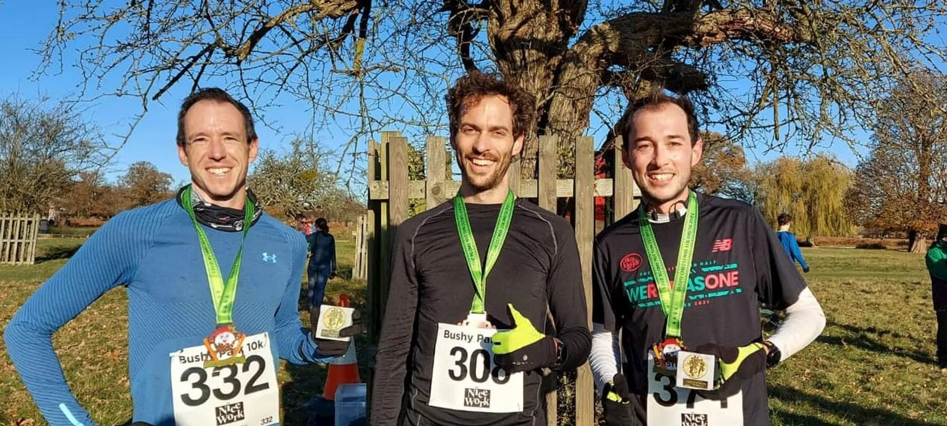 The image shows three individuals posing for a photo outdoors. They are each wearing runner's bibs with numbers (332, 300, 354), indicating they likely just participated in a race, possibly a 10k run given the text on the bib of the person in the middle. They are also wearing finisher medals, suggesting they completed the event. The setting seems to be a park, as indicated by the grass, trees, and the fence in the background. It's a sunny day, and the shadows on the ground suggest it's either morning or late afternoon. Each participant is dressed in athletic wear suitable for running, and they seem to be in good spirits, possibly satisfied with their performance in the race.