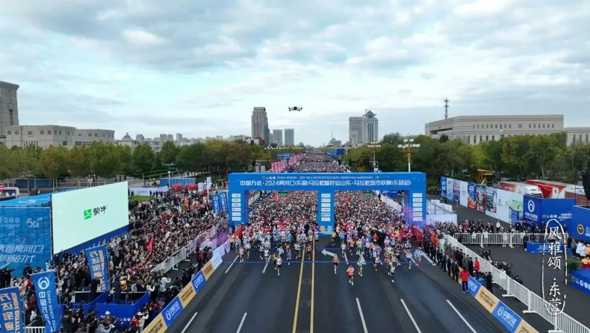 The image shows a large marathon event taking place on a wide street, with numerous runners participating. Spectators are lined up along both sides, and banners stretch across the start line. The scene appears to be festive and well-organized.