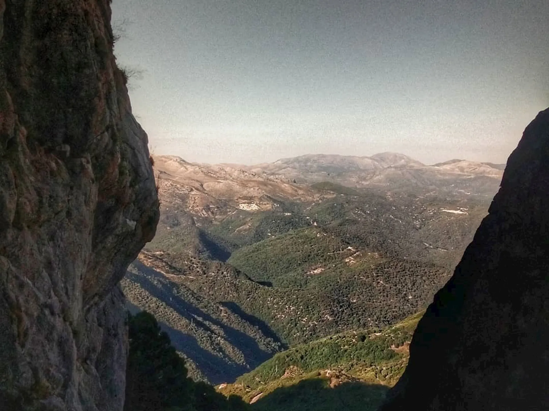 The image shows a scenic landscape viewed from a high vantage point, possibly a mountainside or cliff. We can see a vast expanse of rolling hills or low mountains covered with vegetation, likely trees or shrubs, suggesting a natural, possibly Mediterranean or temperate environment. The foreground on the left is dominated by a rocky outcrop or cliff face that frames the view, leading the eye across the landscape towards the horizon. The sky is clear, indicating good weather conditions, and the lighting suggests it could be either morning or late afternoon. There is no visible human activity or structures, emphasizing the natural and possibly remote character of this location.