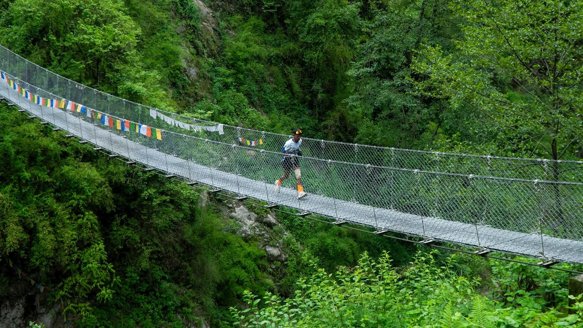 The image shows a person running across a suspension bridge surrounded by lush greenery. The bridge is adorned with colorful prayer flags. The setting is a forested area with abundant vegetation.