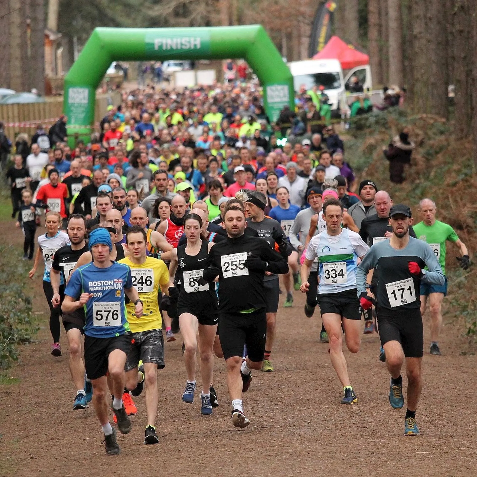 The image shows a group of runners participating in a race. They are approaching or passing under a green inflatable arch that reads "FINISH," indicating it is the end of the race. The runners are wearing athletic clothing and race bibs with numbers, which are typically worn during competitive events to identify participants. The environment suggests the race is taking place in a park or wooded area with trees lining the path. Some spectators and event organizers appear to be just beyond the finish line, although they are not the main focus of the image. The runners have various expressions, which often reflect a mix of exhaustion, determination, and accomplishment as they complete the race.
