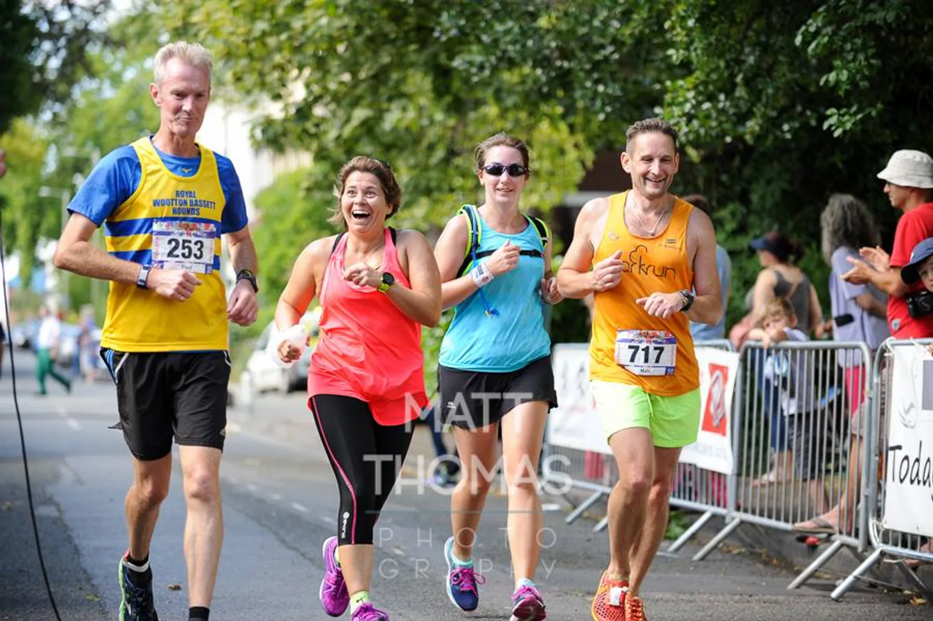 The image shows four people participating in a road race. All of them are wearing bib numbers, which suggests that they are competitors in an organized running event, such as a marathon, half-marathon, or shorter road race. They appear to be enjoying themselves, with one woman in the center smiling and laughing. All runners are captured in mid-stride, indicating the dynamic nature of the event. There are spectators on the right-hand side, watching and cheering from behind barriers. The environment is a tree-lined street, suggestive of an urban or park setting. The watermark suggests that this photo was taken by a professional photographer, likely for the purpose of selling the photos to the runners as a keepsake from the event.