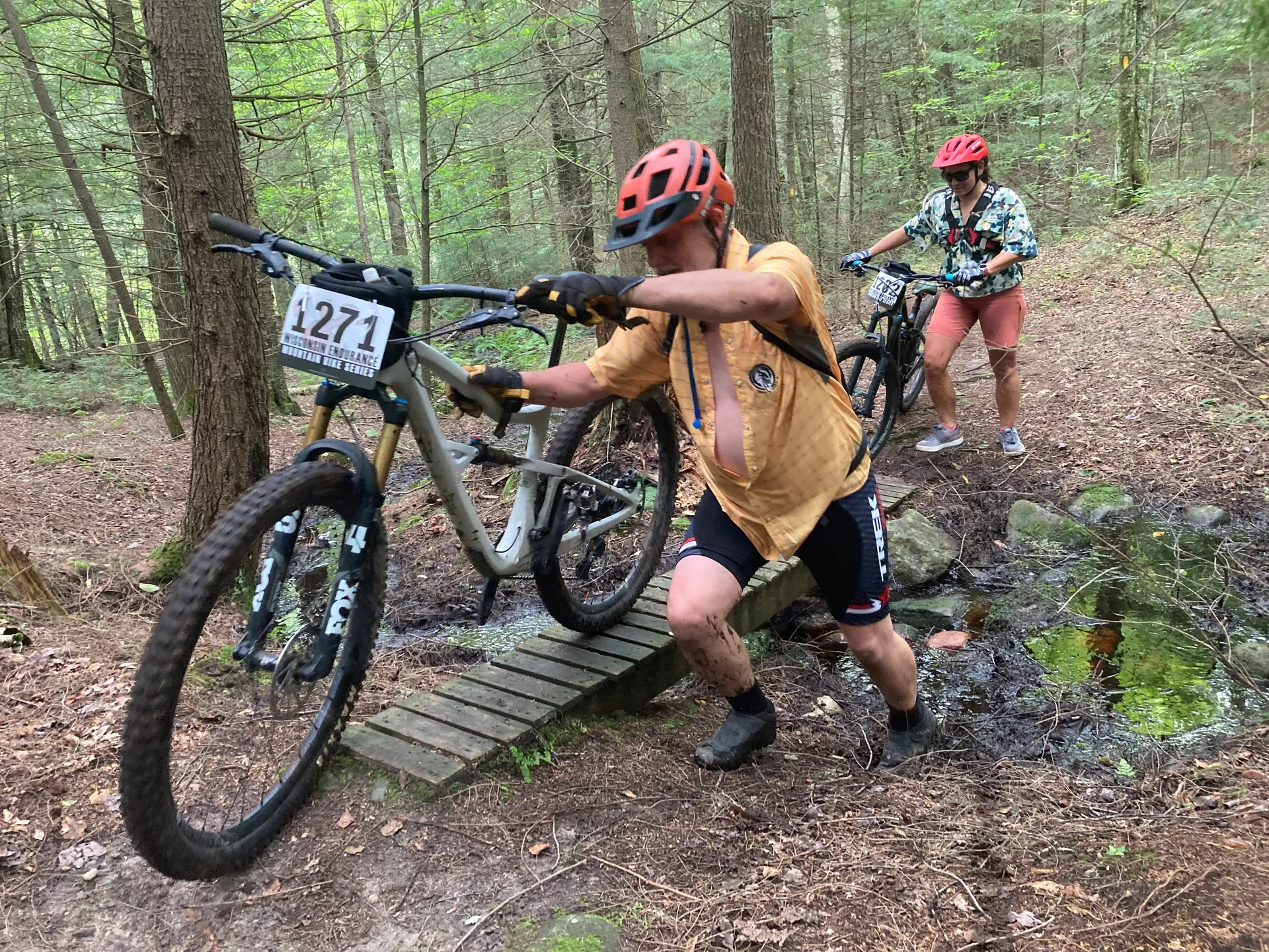 The image shows two people with bicycles in a forest setting. They are navigating a small bridge or wooden plank over a muddy or uneven area. The person in front is pushing their bicycle up the bridge, while the person behind is walking with their bicycle as well. Both individuals are wearing helmets.