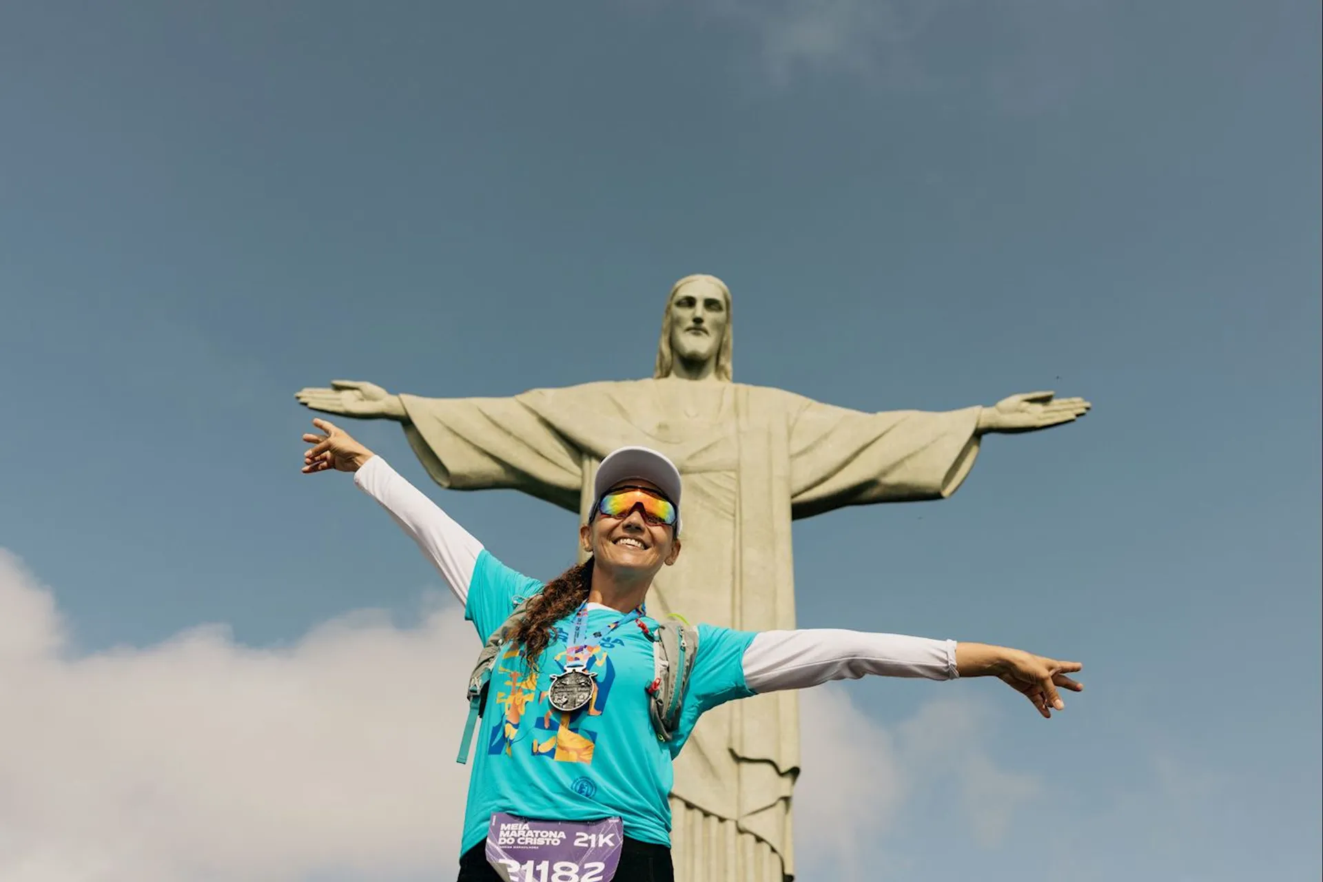 The image shows a person posing with their arms outstretched in front of the Christ the Redeemer statue in Rio de Janeiro, Brazil. The person is wearing a cap, sunglasses, a blue race bib, and has medals around their neck.