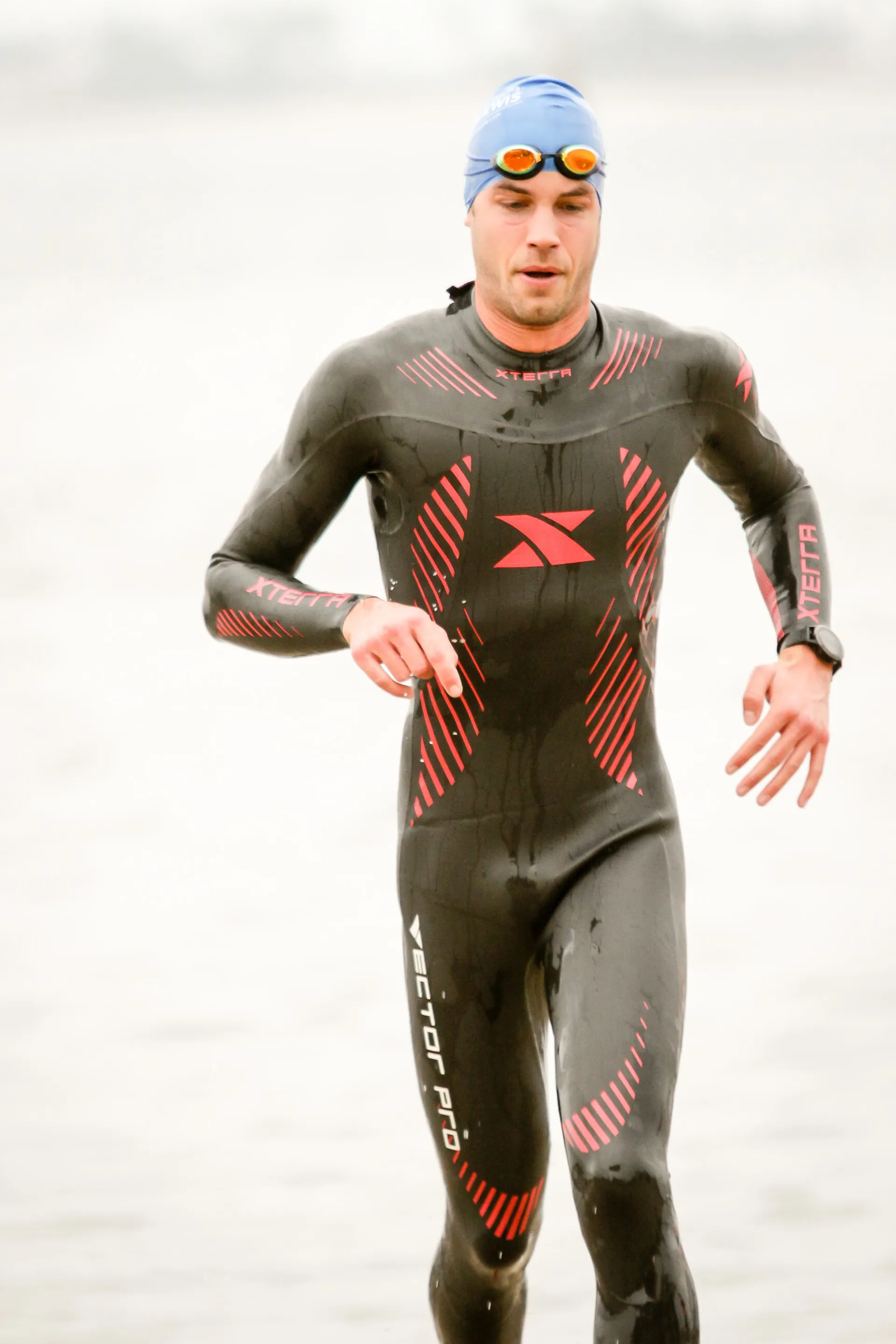 The image shows a person wearing a wetsuit, swim cap, and goggles. The wetsuit is black with red designs, which suggests it is designed for activities such as swimming, triathlon, or water sports where thermal insulation and streamlining are beneficial. The person appears to be in motion, possibly running or walking briskly, which could indicate they are participating in a triathlon or open water swimming event. The background is quite overexposed with a diffuse, light color, making it difficult to discern the exact environment, but it suggests a bright, possibly outdoor setting.