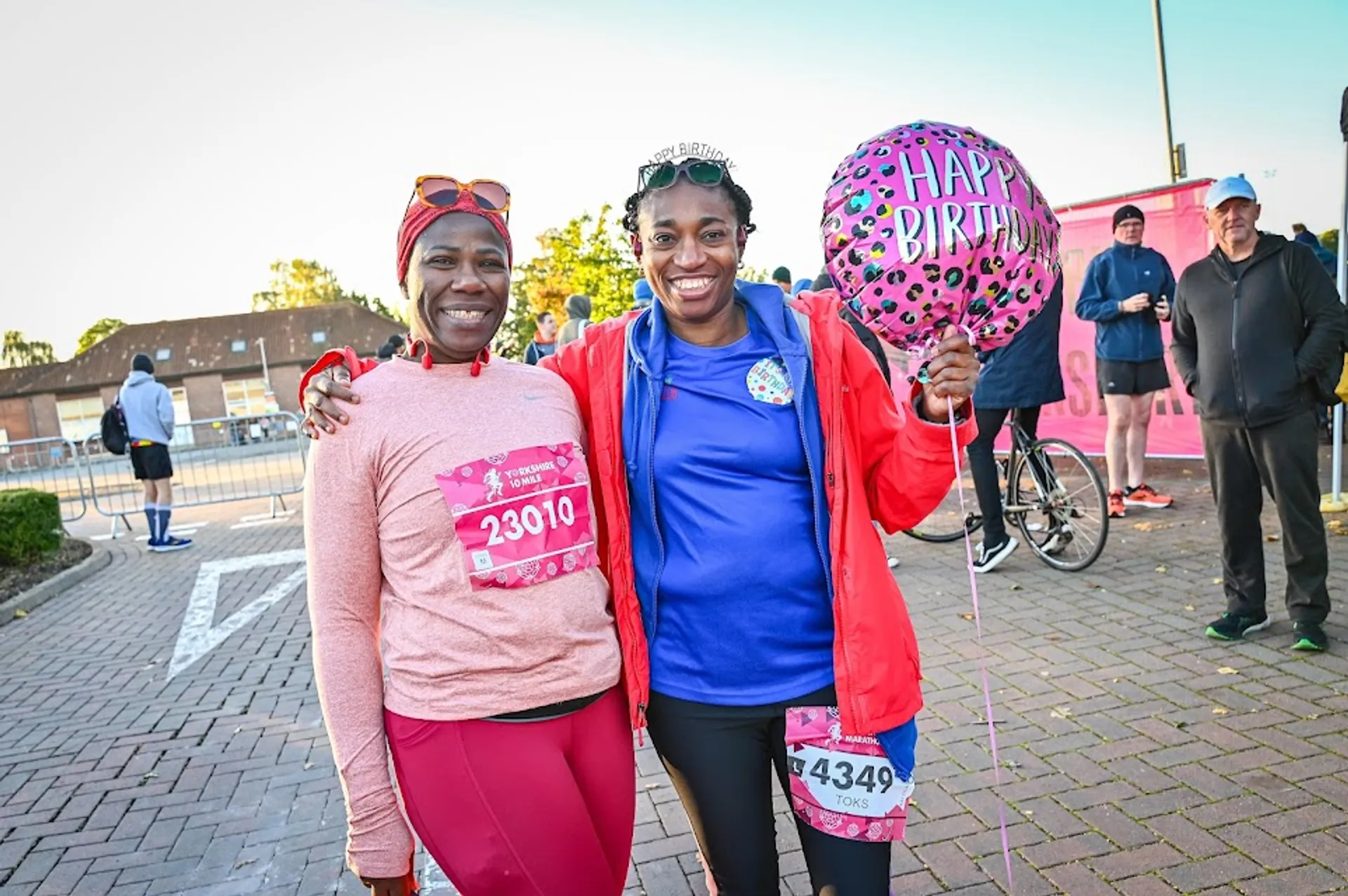 In the image, there are two women standing side by side, smiling at the camera. They seem to be at some sort of outdoor event, possibly a race given the numbers pinned to their shirts (23010 and 43449). The woman on the right is wearing a tiara and holding a balloon with the words "Happy Birthday" on it, indicating that it may be her birthday. Both are dressed in athletic clothing suitable for running, with the dominant colors being pink and blue. There are other individuals and structures in the background which suggest that this is a public space, such as a park or event plaza.