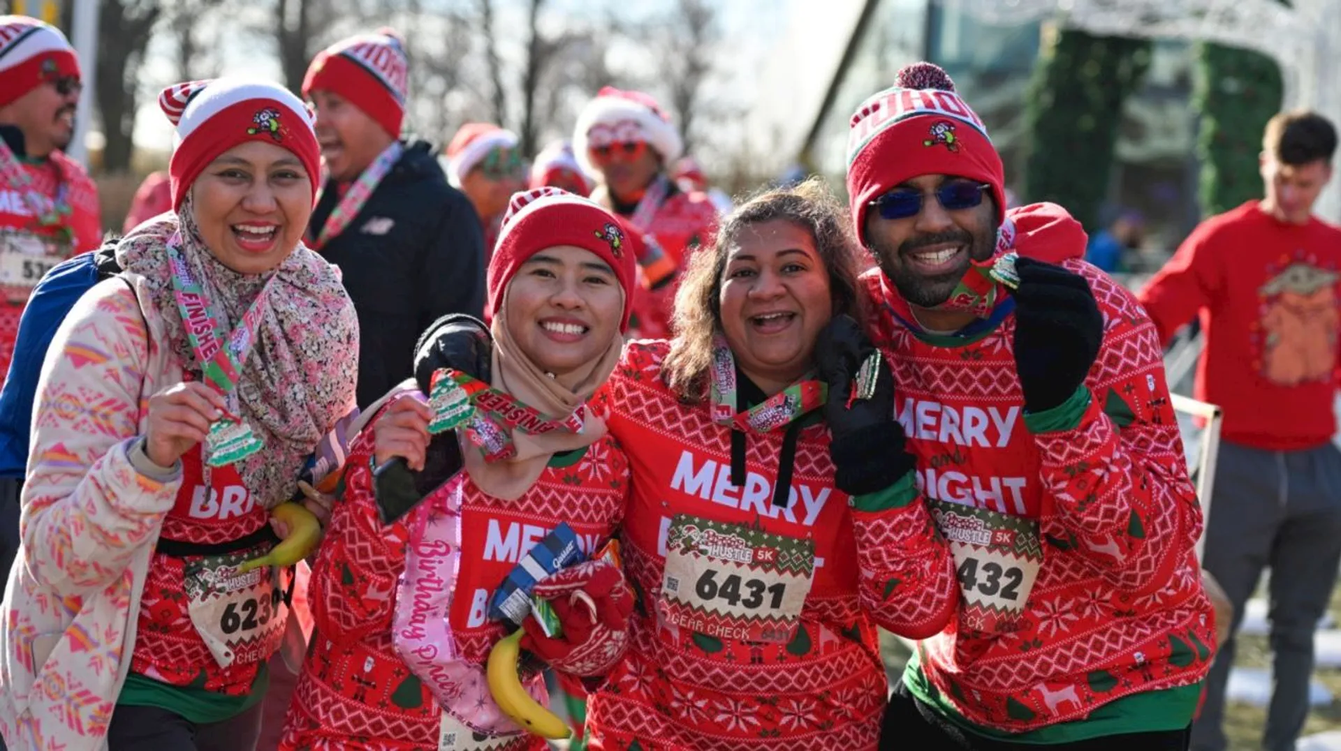 The image shows a group of people participating in a festive run, likely themed around Christmas, given their holiday sweaters and Santa hats. They appear to be celebrating, possibly having completed the event, as they are holding medals and looking cheerful.