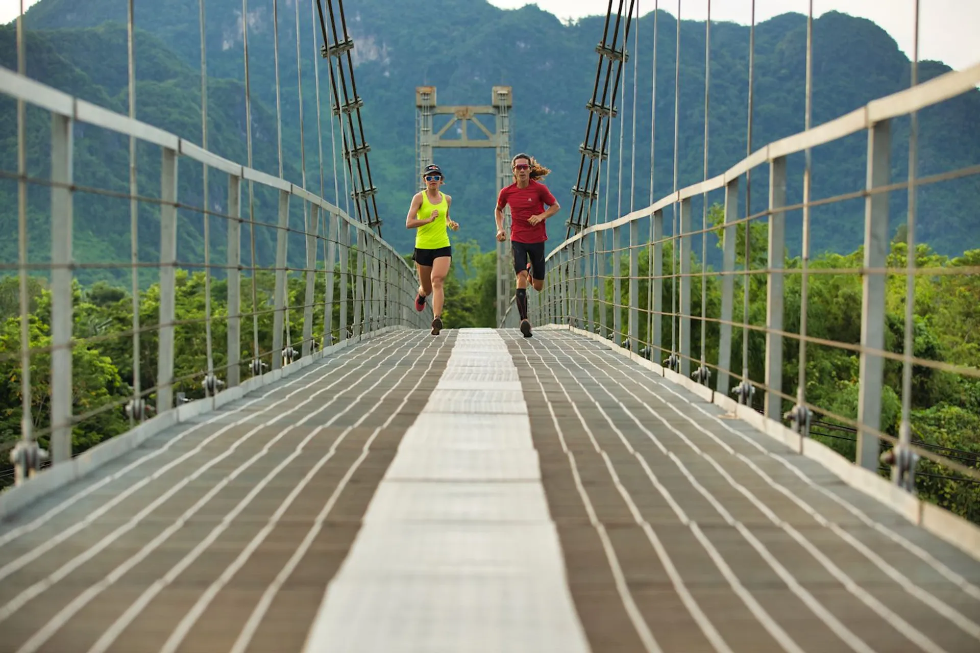 The image shows two people jogging towards the camera on a suspension bridge. The bridge